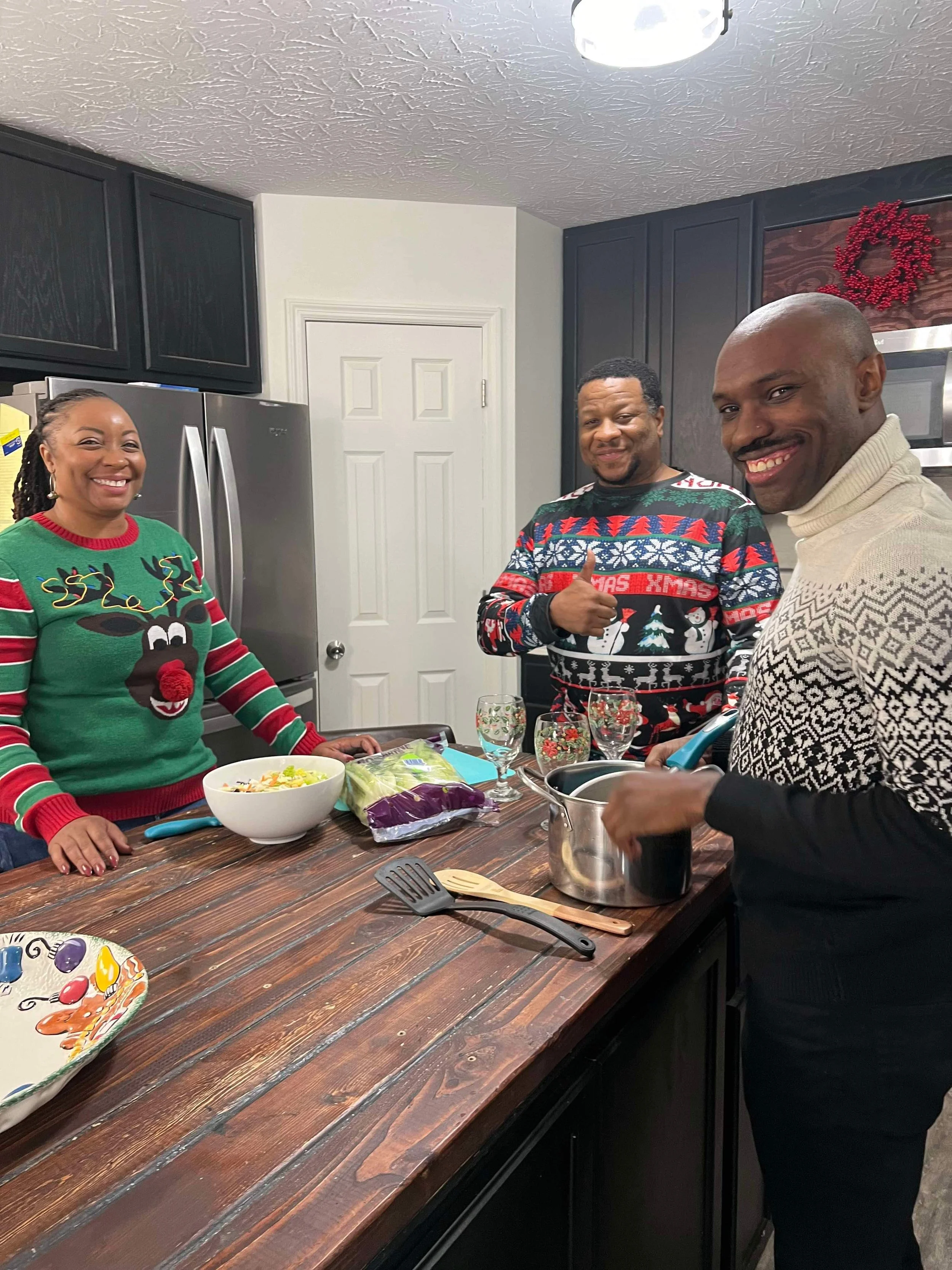 Three people in Christmas sweaters in a kitchen smiling at the camera, with a wooden kitchen island, a bowl of salad, vegetables, and cooking utensils on the counter.