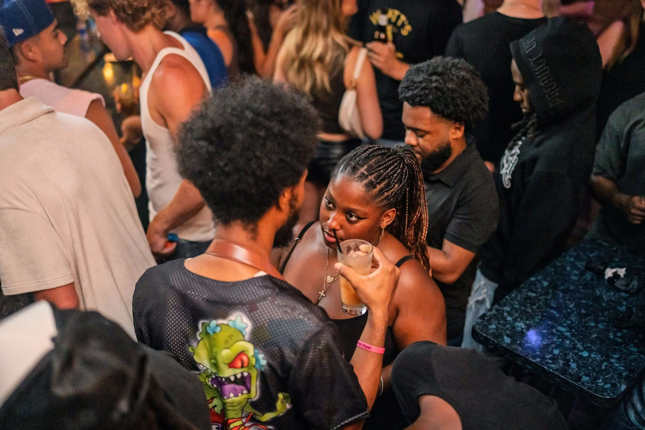 A group of people socializing at a crowded indoor event, with two women in the foreground engaged in conversation, one holding a drink.