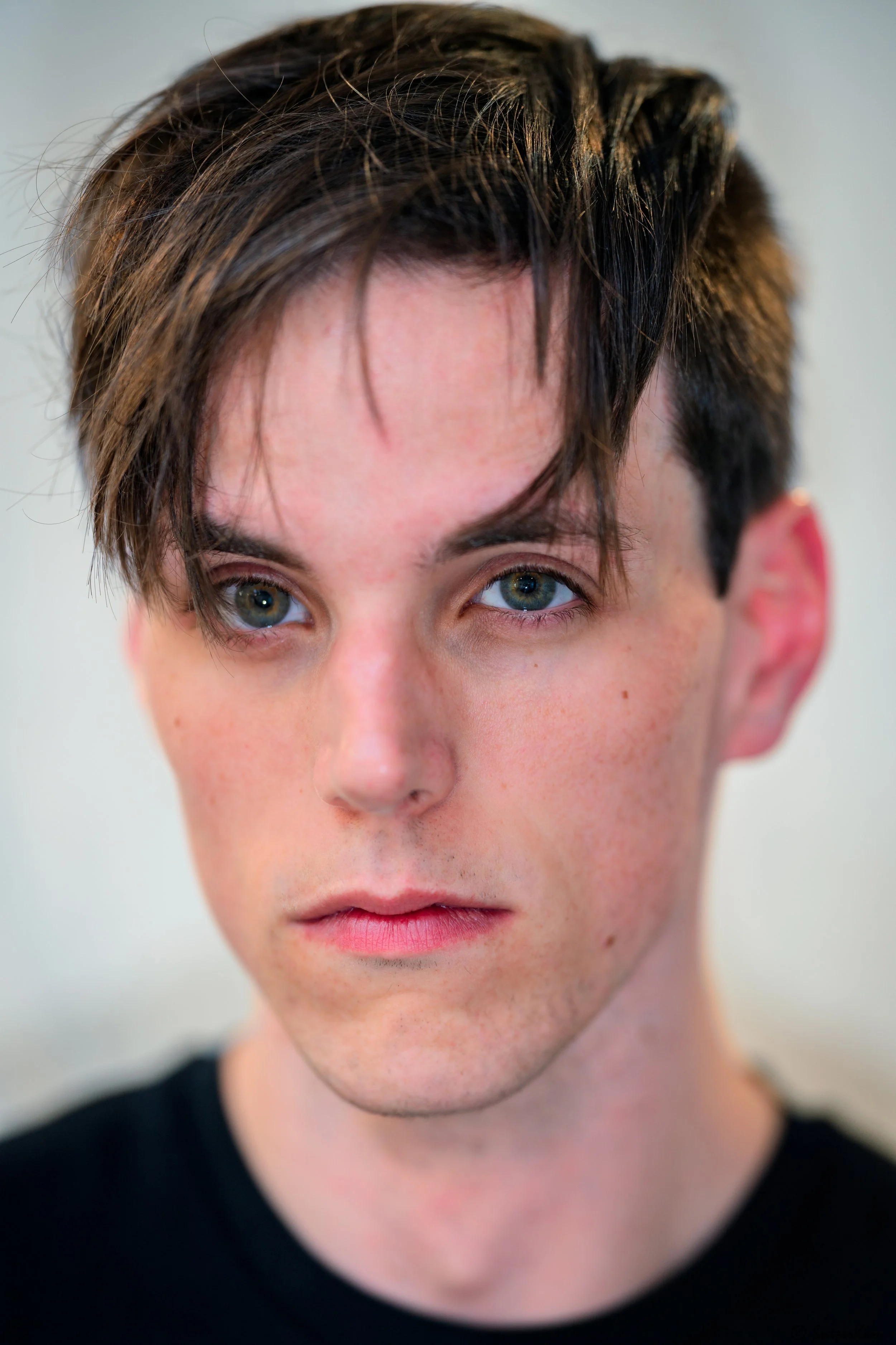 Close-up portrait of a young man with blue eyes and dark brown hair, wearing a black shirt, looking directly at the camera.