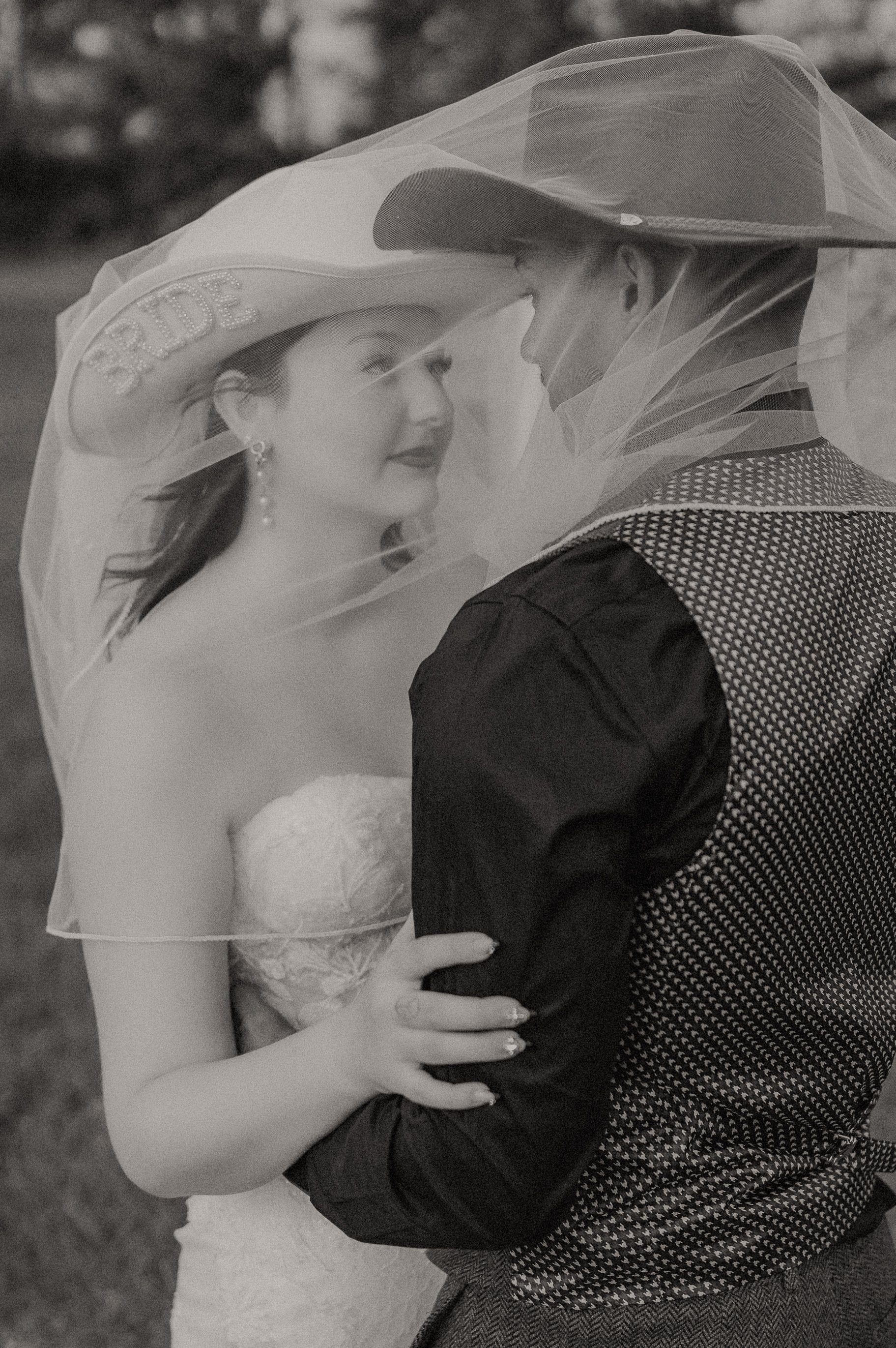 A bride and groom standing closely outdoors, with the bride wearing a cowboy hat with 'BRIDE' written on it and a veil, the bride has earrings and is in a strapless dress, and the groom is in a patterned vest over a shirt, both looking at each other.