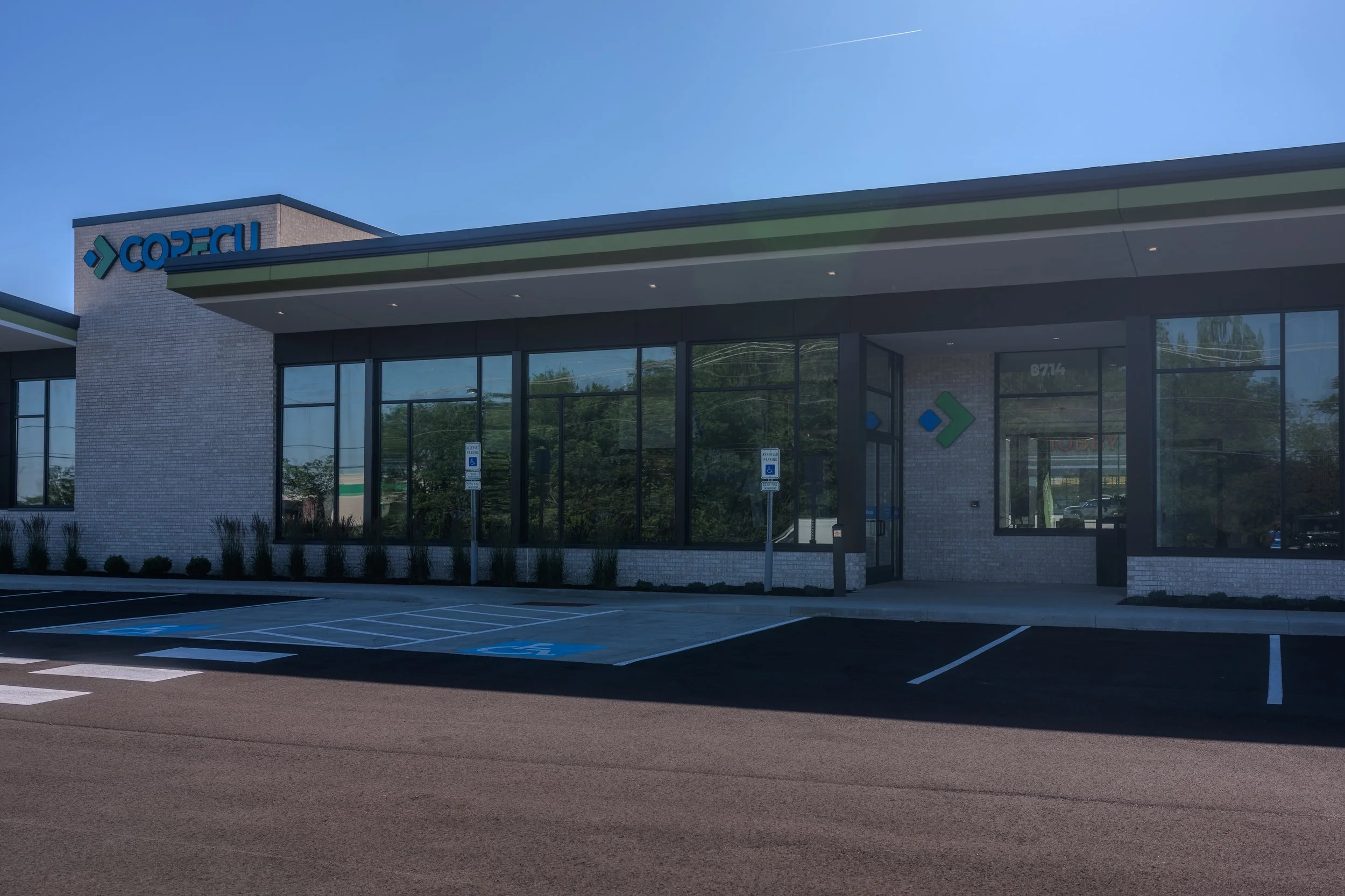 A modern credit union with a sign that reads 'COPFCU' on the top corner. The building has large glass windows and parking spots in front, including designated handicapped spaces. The sky is clear and blue.