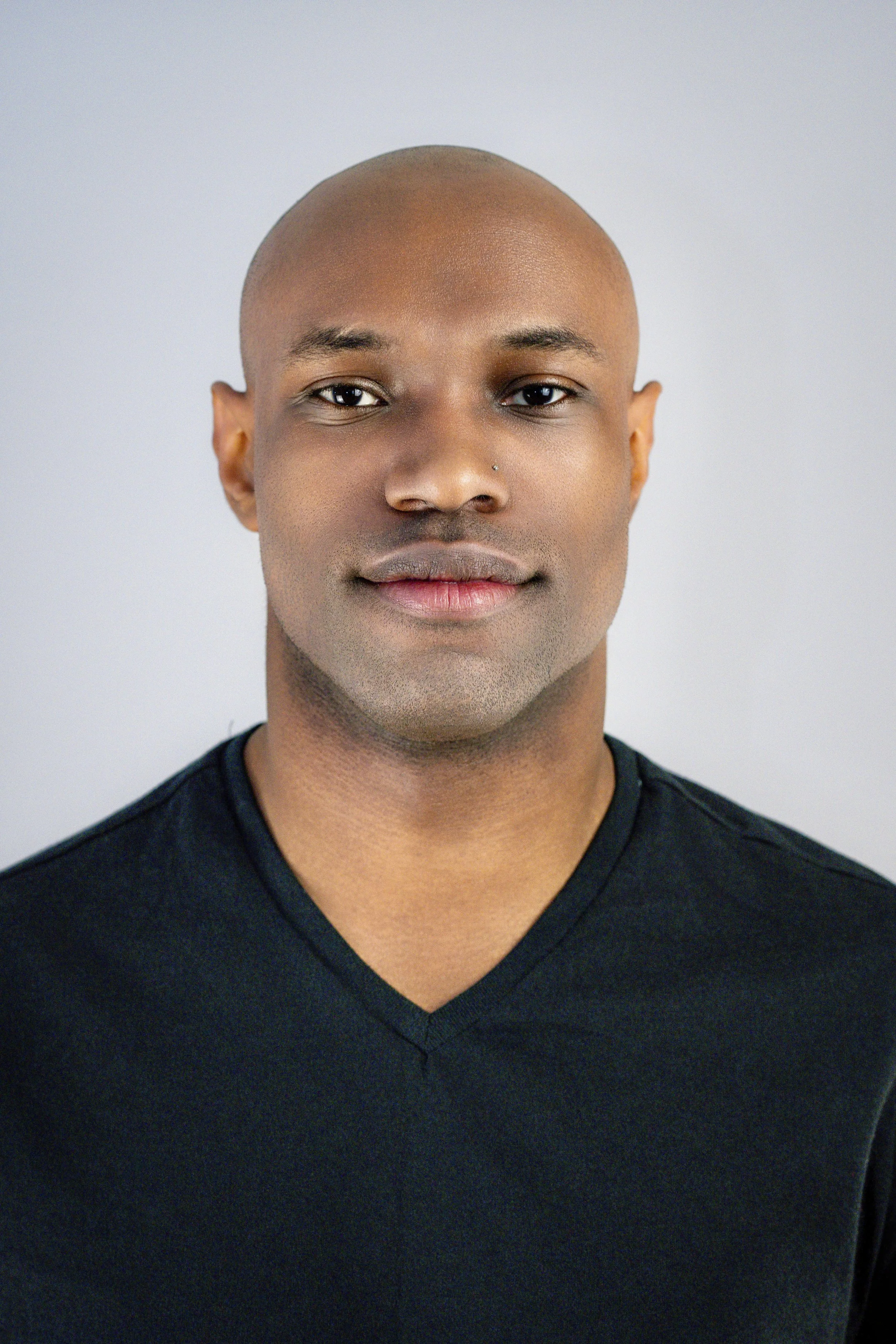 A portrait of a man with a bald head, dark complexion, and wearing a black V-neck shirt, against a plain light gray background.