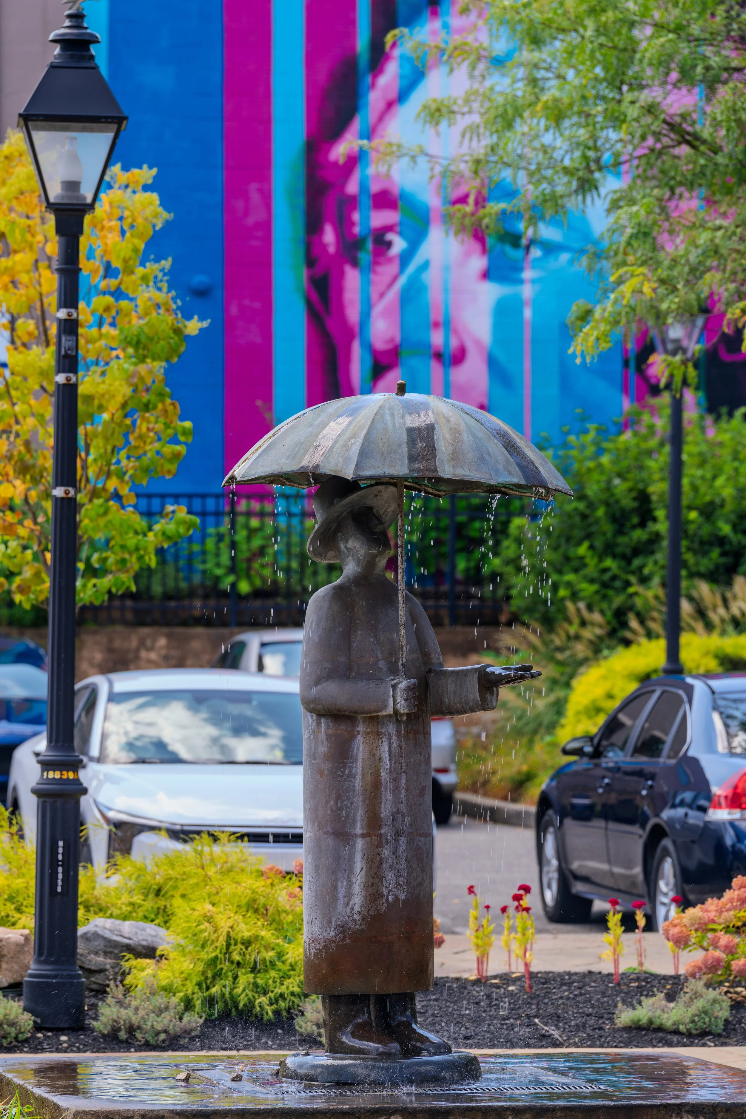 Bronze statue of a person holding an umbrella, with water flowing off it, standing in a landscaped area with a colorful mural of a woman's face in the background, trees, and parked cars.