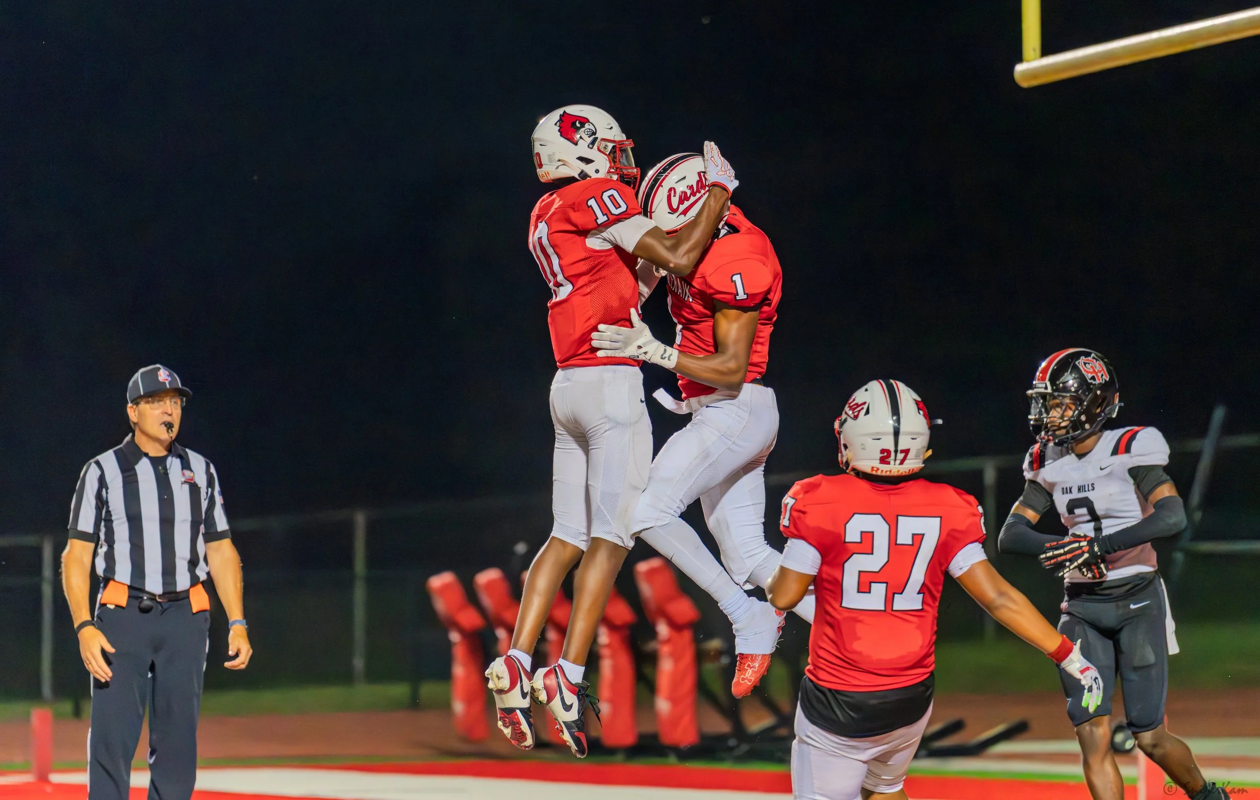 High school football players in red and black uniforms celebrate a touchdown on the field at night, with an official watching nearby.