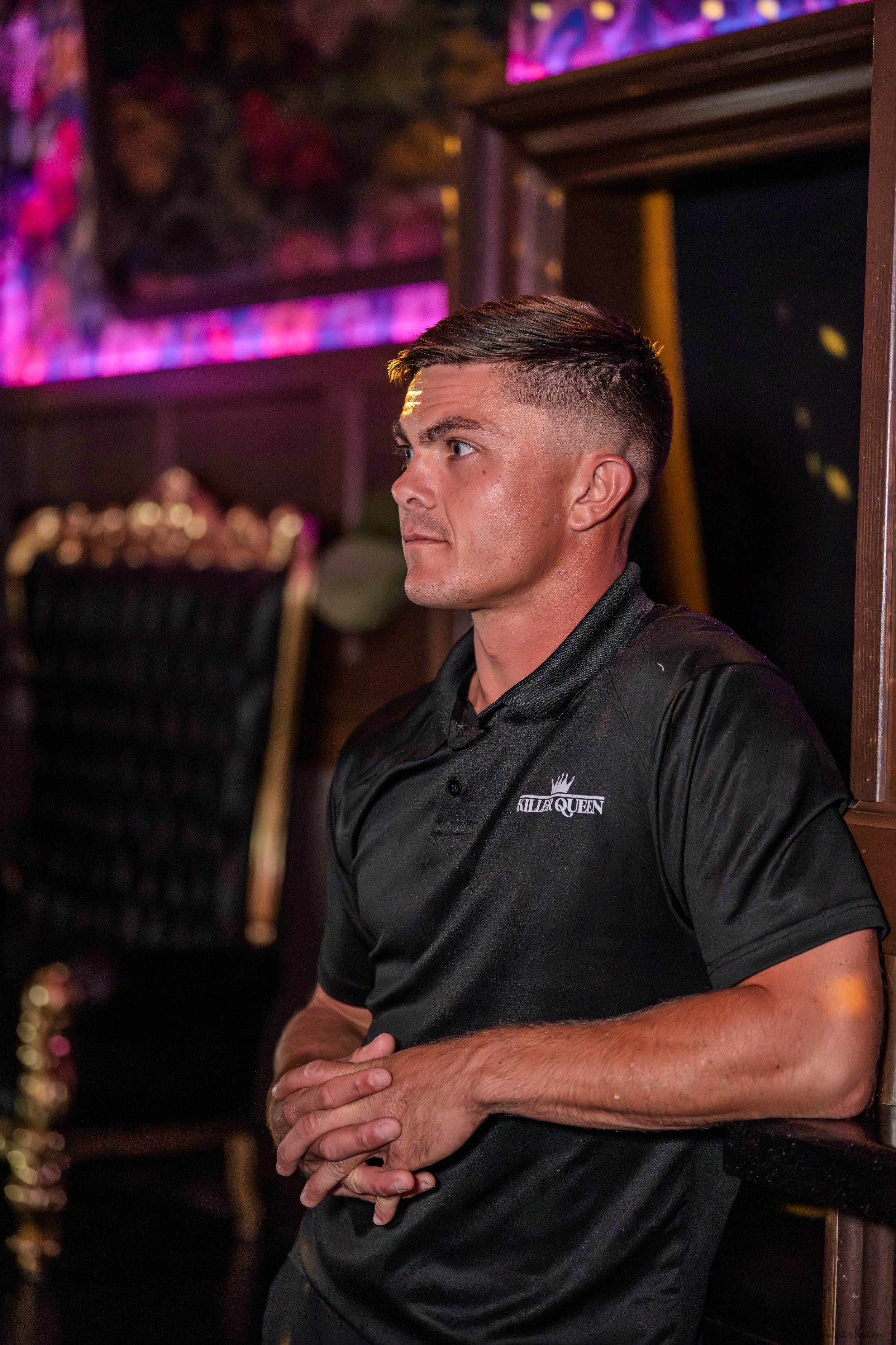 A young man with short brown hair styled in a fade, wearing a black polo shirt with the logo 'KILLA QUEEN' on the left chest, stands indoors with his arms crossed, looking to the left. The background is dimly lit with purple and pink decorative lighting.