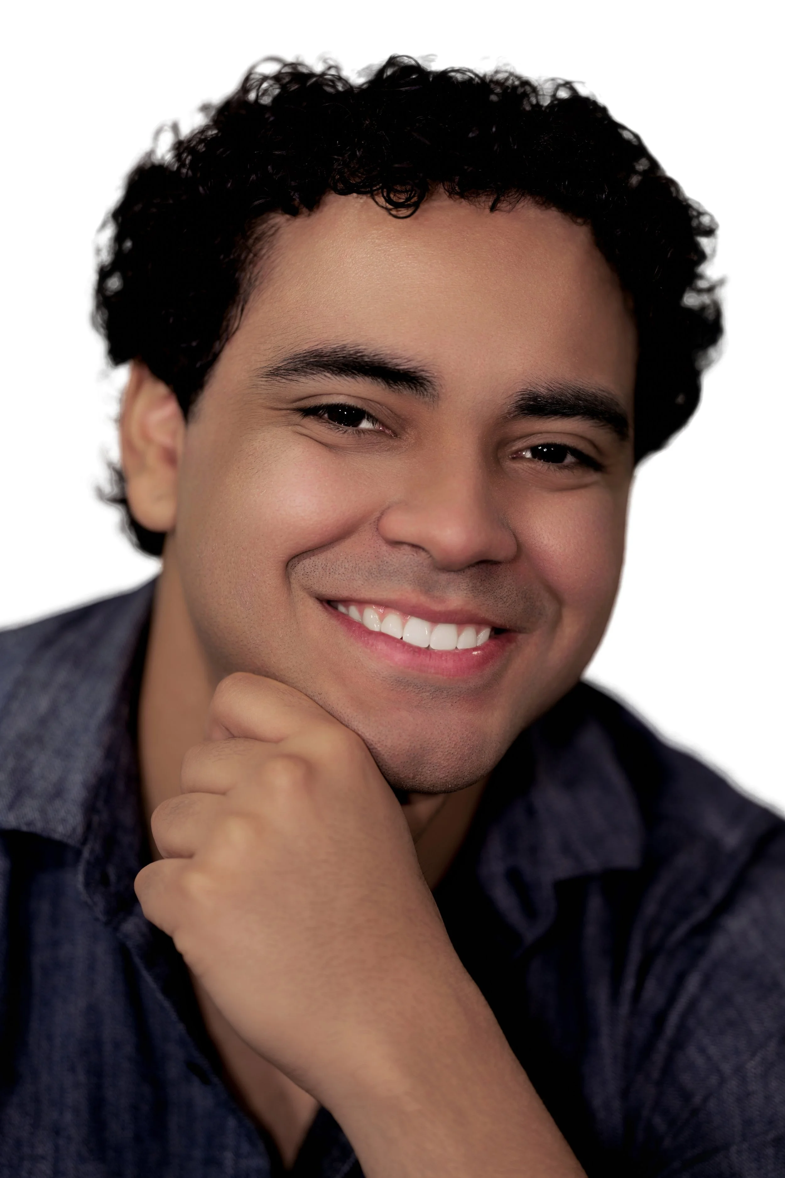 Close-up portrait of a smiling young man with curly dark hair, wearing a dark denim shirt, resting his chin on his hand against a plain white background.