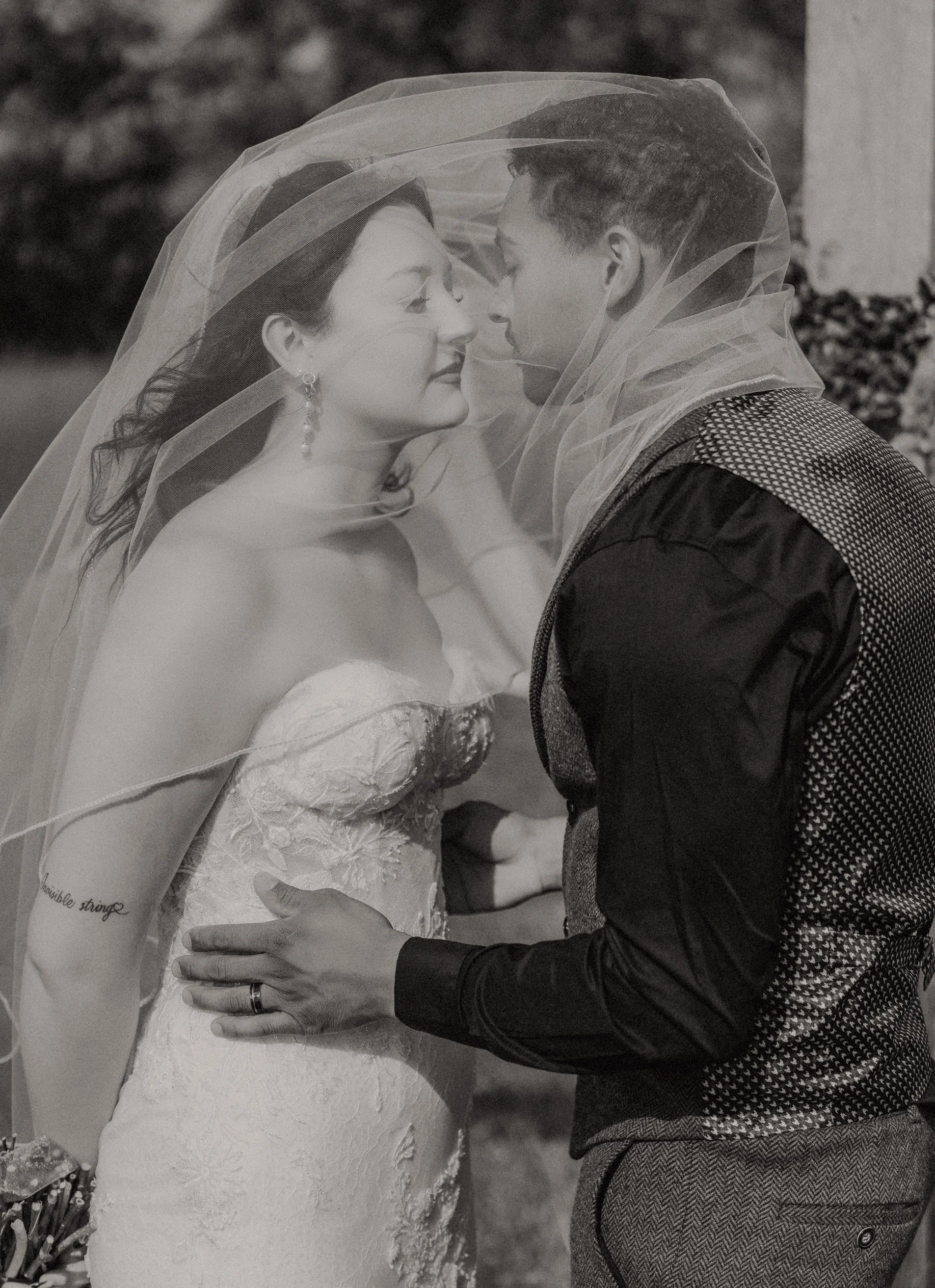 A bride and groom sharing a close, intimate moment outdoors, with their faces nearly touching and veils draped over their heads in a black and white photograph.
