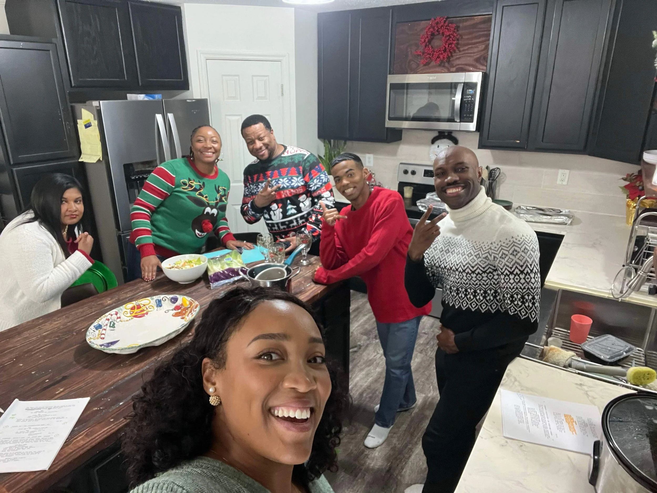 A group of six people in a kitchen celebrating Christmas, wearing festive sweaters, smiling, and making peace signs. The kitchen has black cabinets, a stainless steel refrigerator, microwave, and holiday decorations.