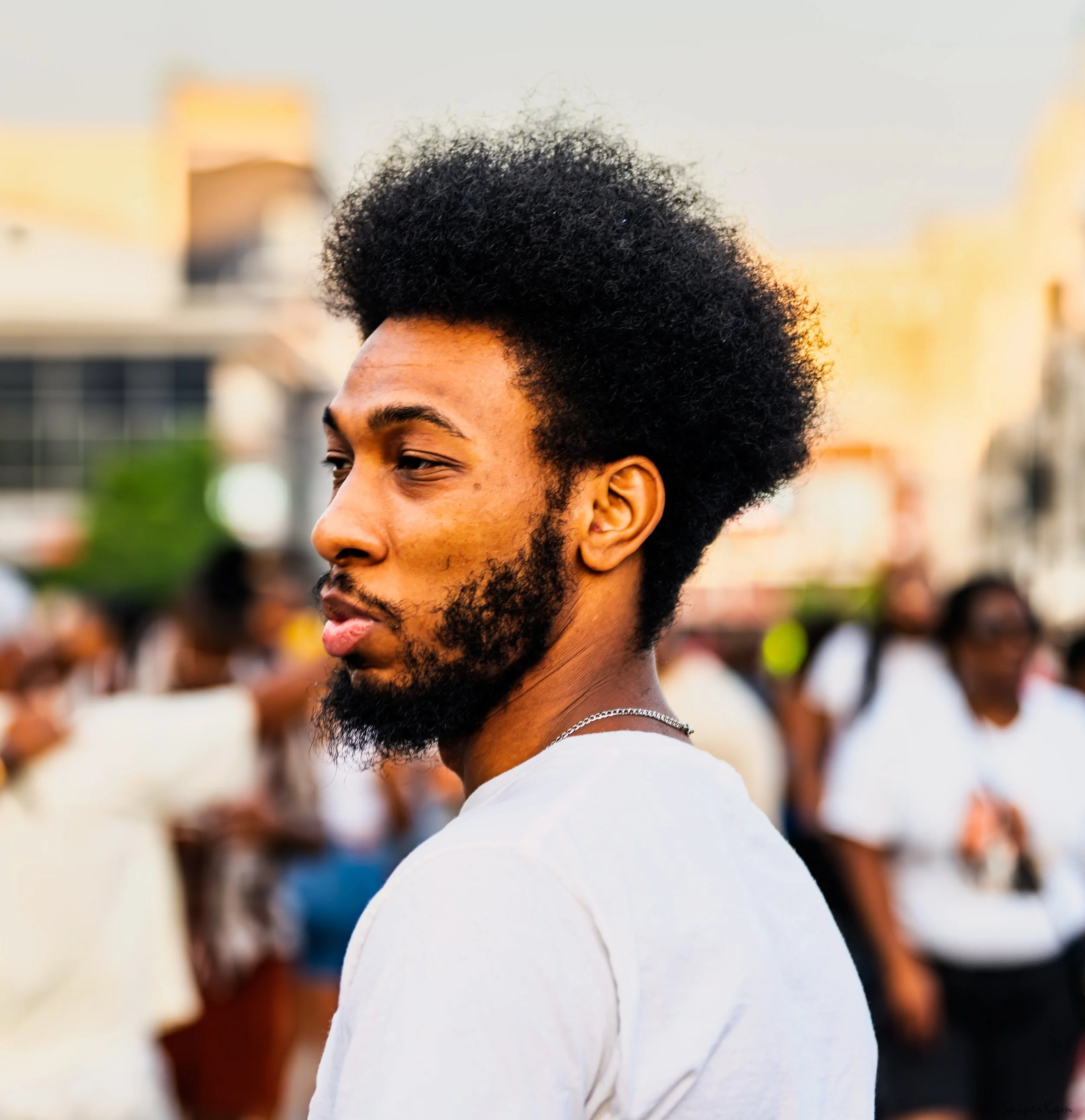 A young man with an afro hairstyle and a beard, wearing a white t-shirt, standing outdoors among a crowd, with buildings and trees in the background, during the evening.