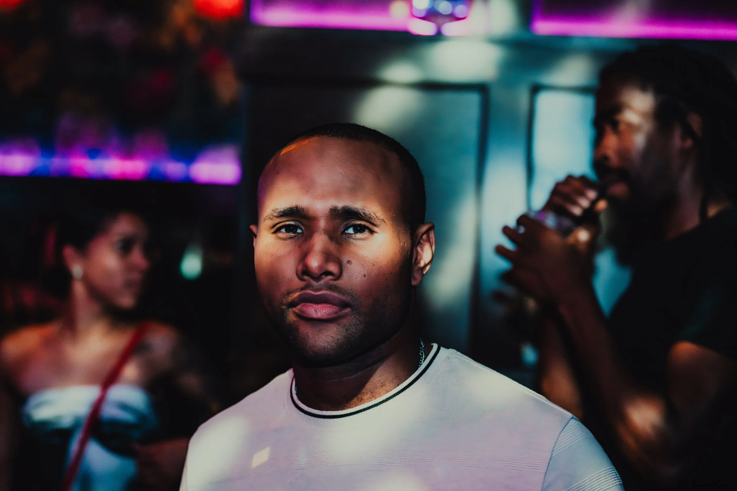 A young man with short dark hair and a serious expression in a dimly lit nightclub or party, with colorful neon lights and blurred figures in the background.