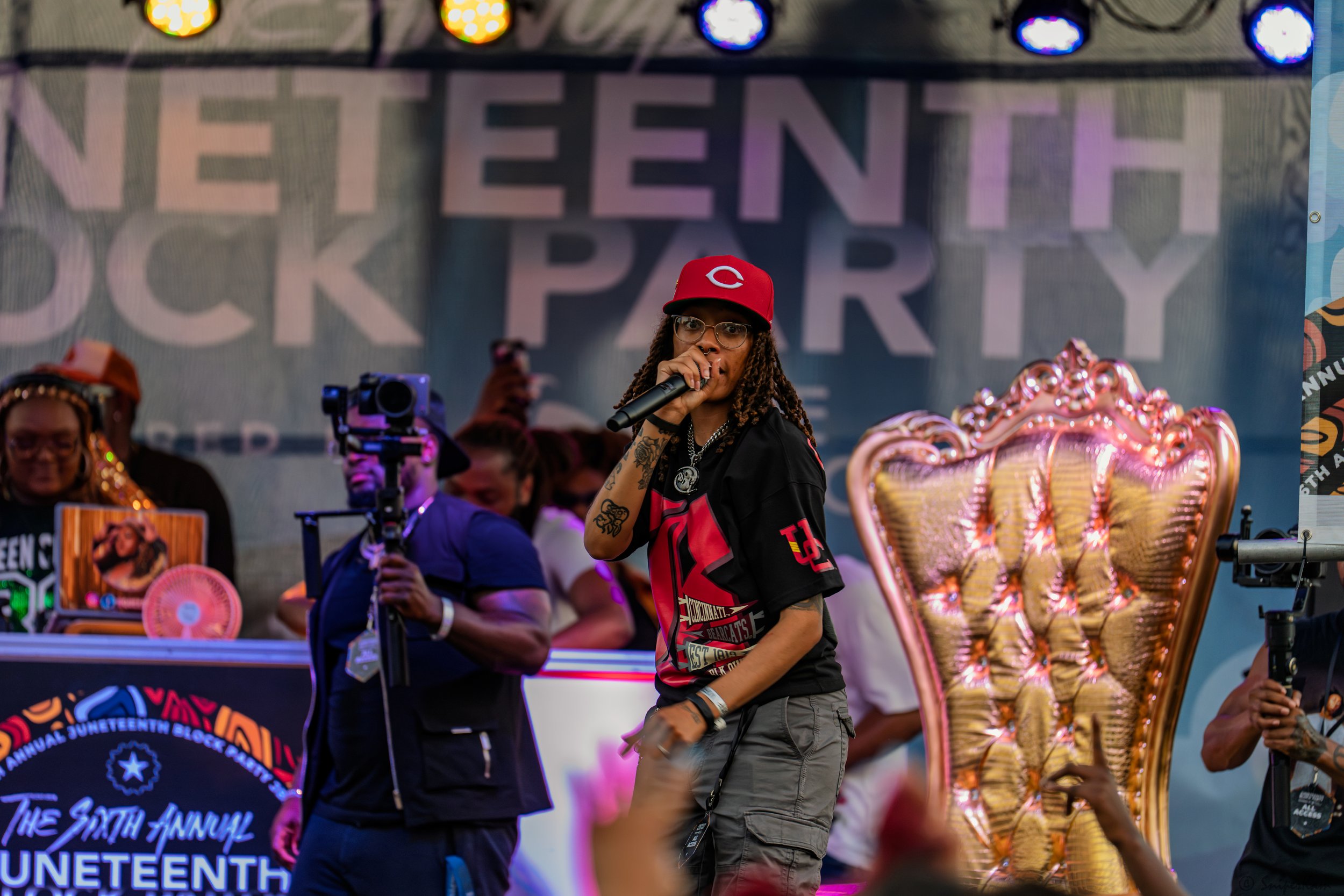 A woman with long dreadlocks, glasses, and tattoos, wearing a red baseball cap and a black sports jersey, holding a microphone on stage at an event with a large backdrop that reads 'Juneteenth Park.' There are people around her, including a man with 