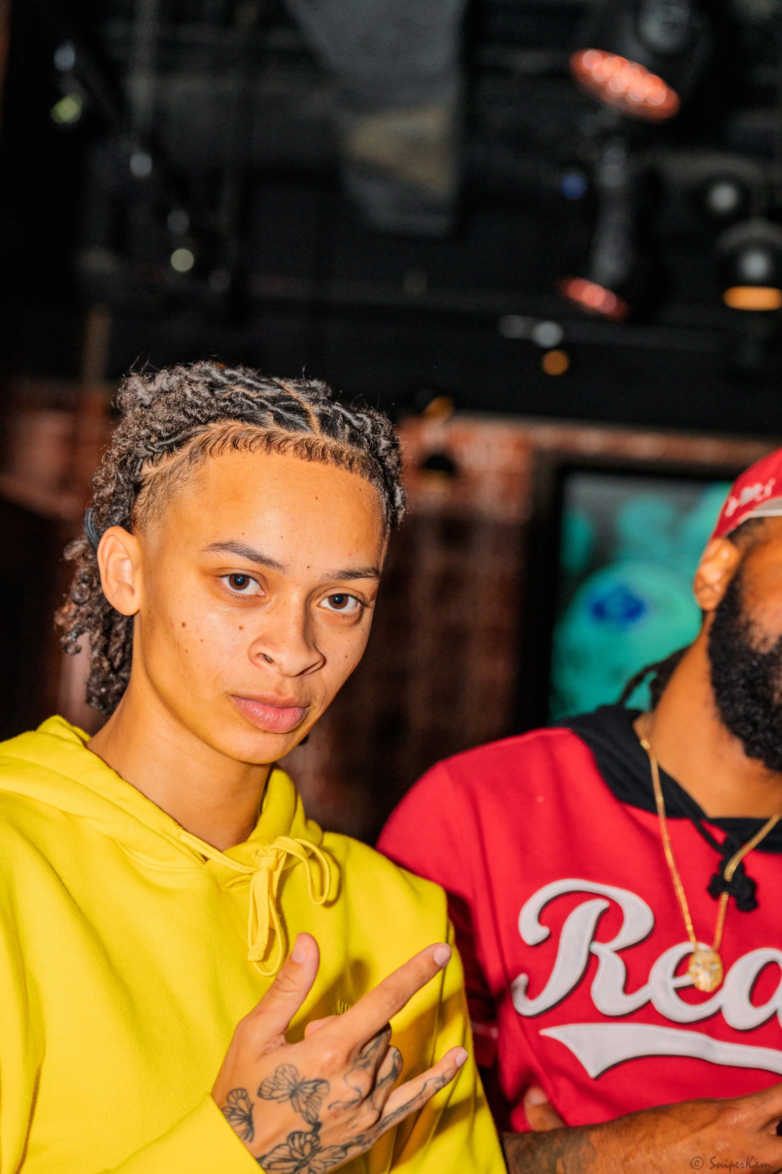 Young woman with braided hair and tattoos on her fingers, wearing a bright yellow hoodie, making a peace sign with her hand, standing next to a person in a red shirt with 