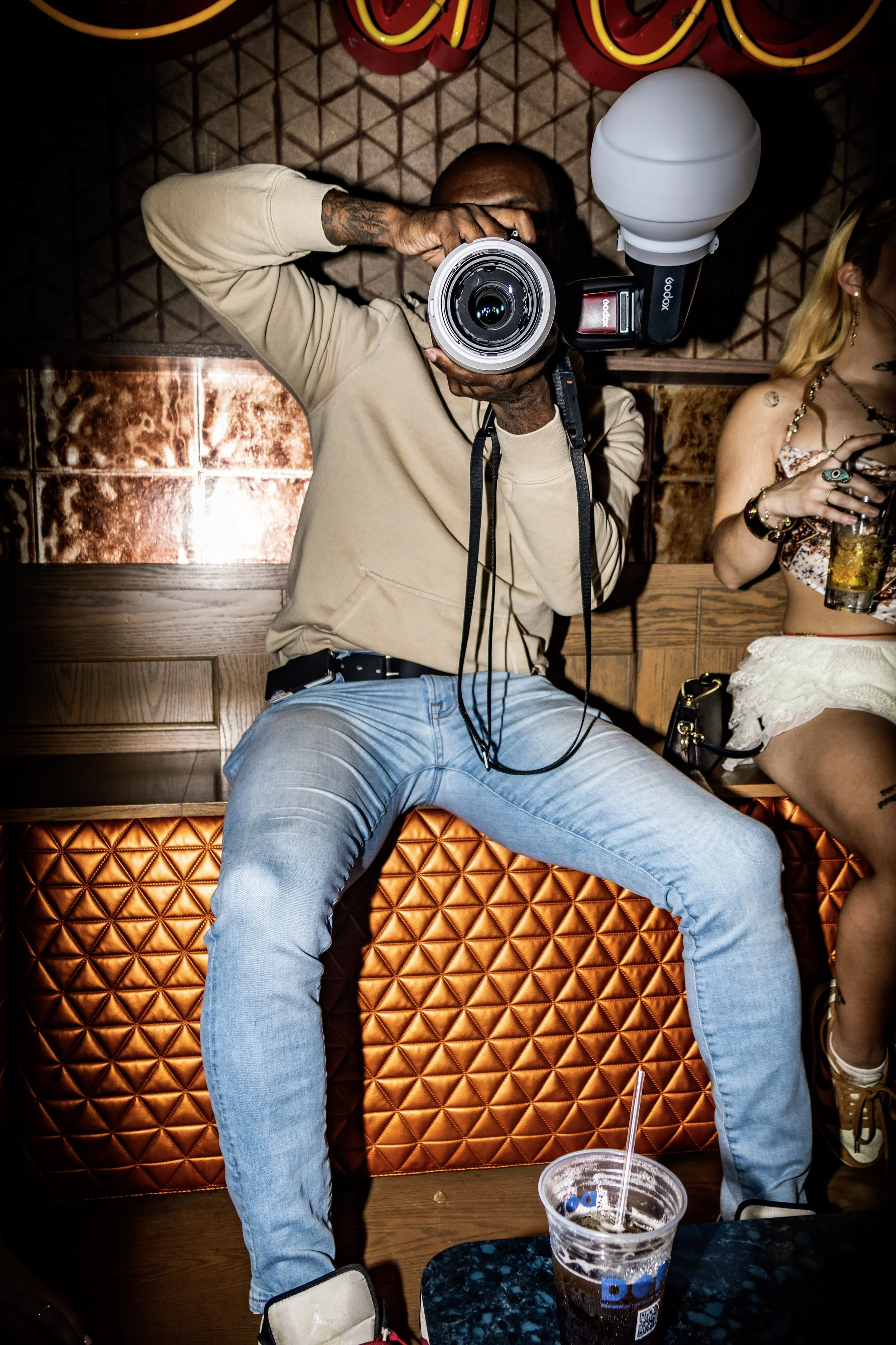 A man holding a camera takes a photo in a bar or lounge with a tatami wall, a neon sign overhead, and a woman sitting next to him holding a drink.