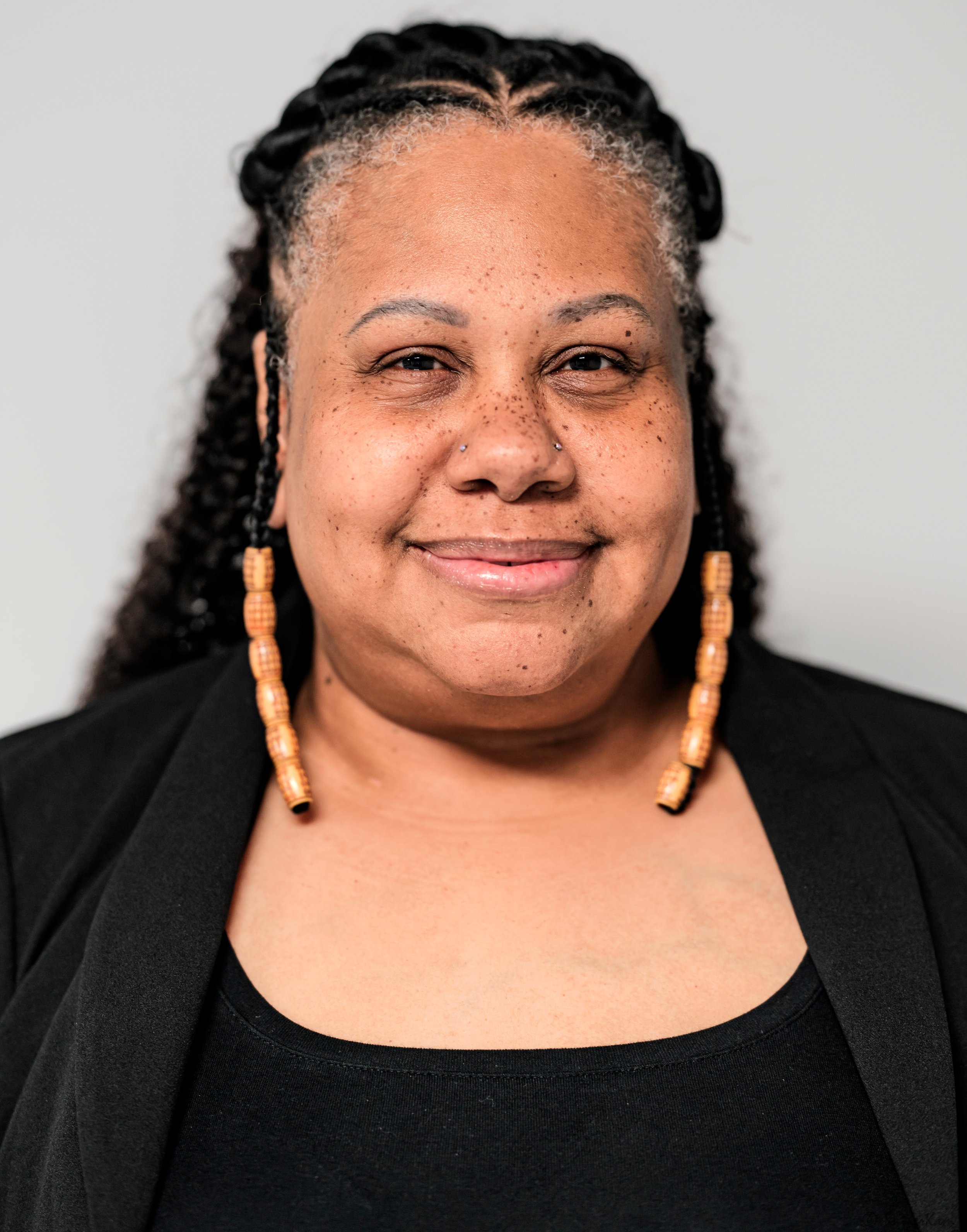 A woman with braided hair, tan beaded earrings, and a smile, wearing a black top and blazer.