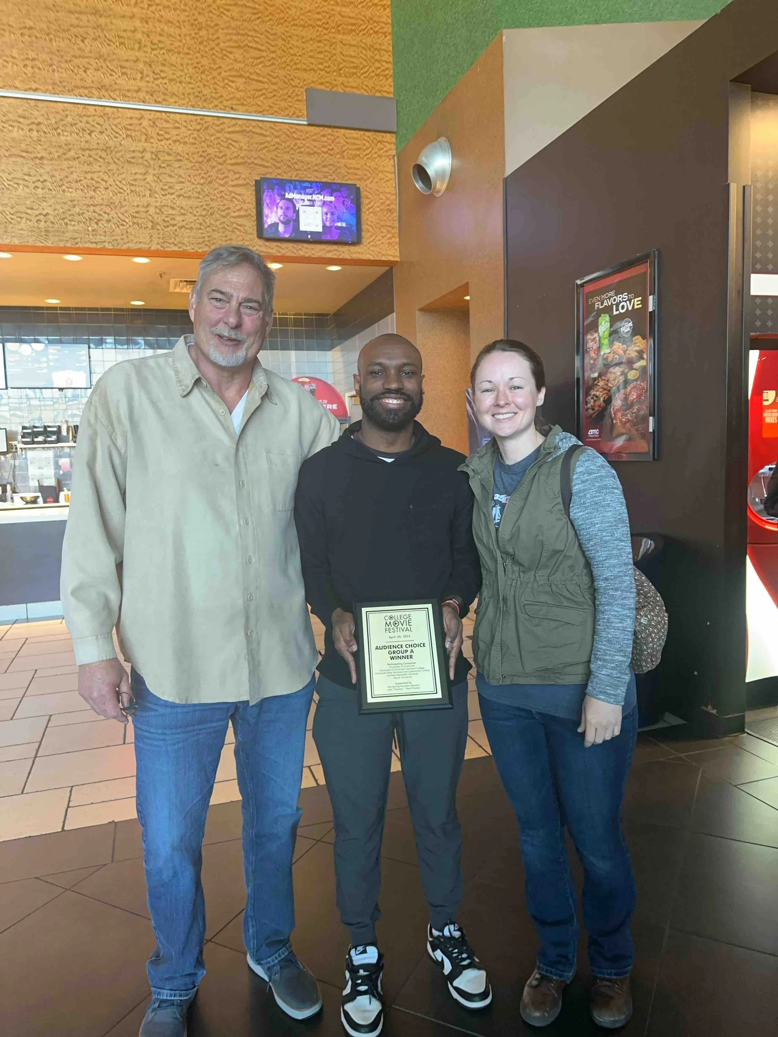 Three people standing side by side inside a movie theater lobby, with the person in the middle holding a certificate, all smiling for the camera.