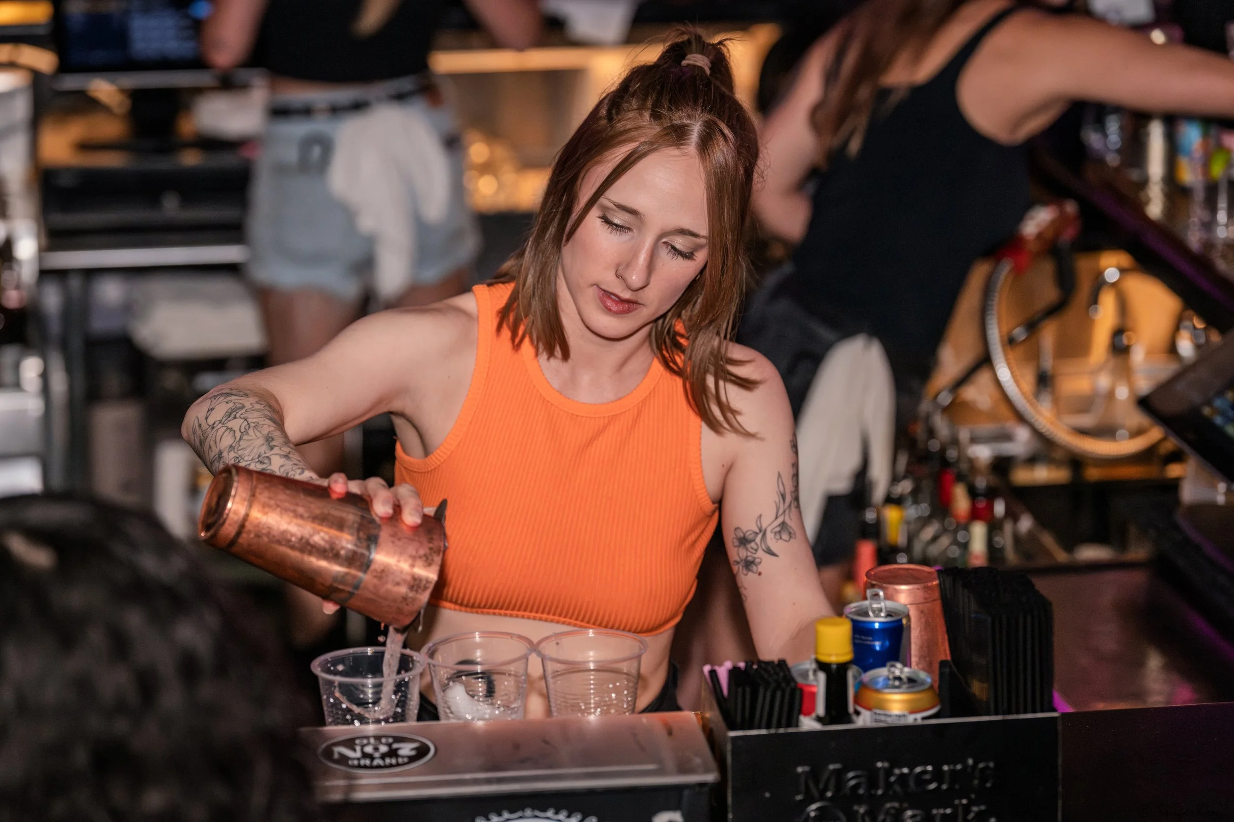 A female bartender with tattoos on her arms, wearing an orange sleeveless top, pours liquid from a copper cocktail shaker into a glass at a bar.