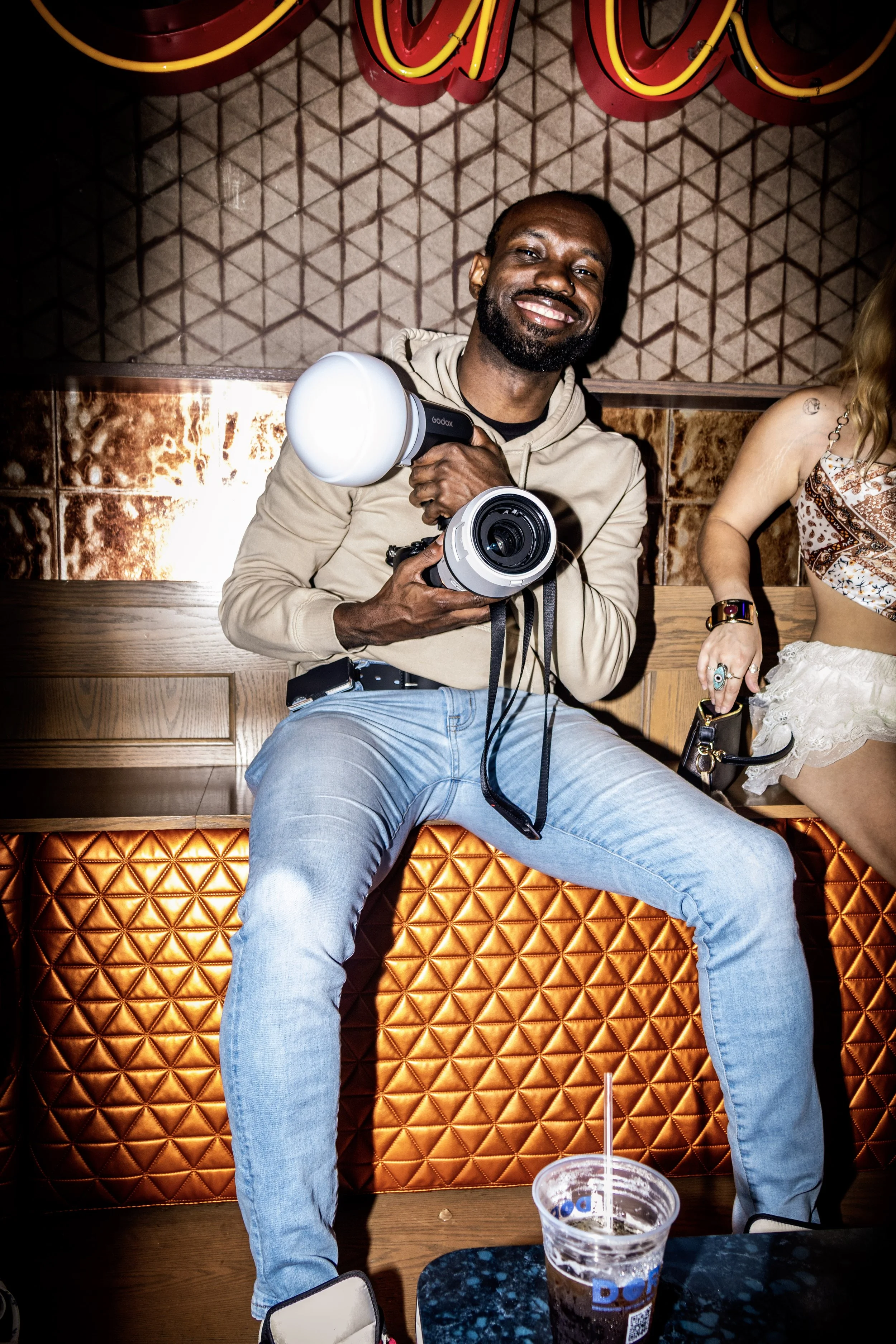 A man sitting on an orange padded bench holding a professional camera with a flash accessory, smiling at the camera.