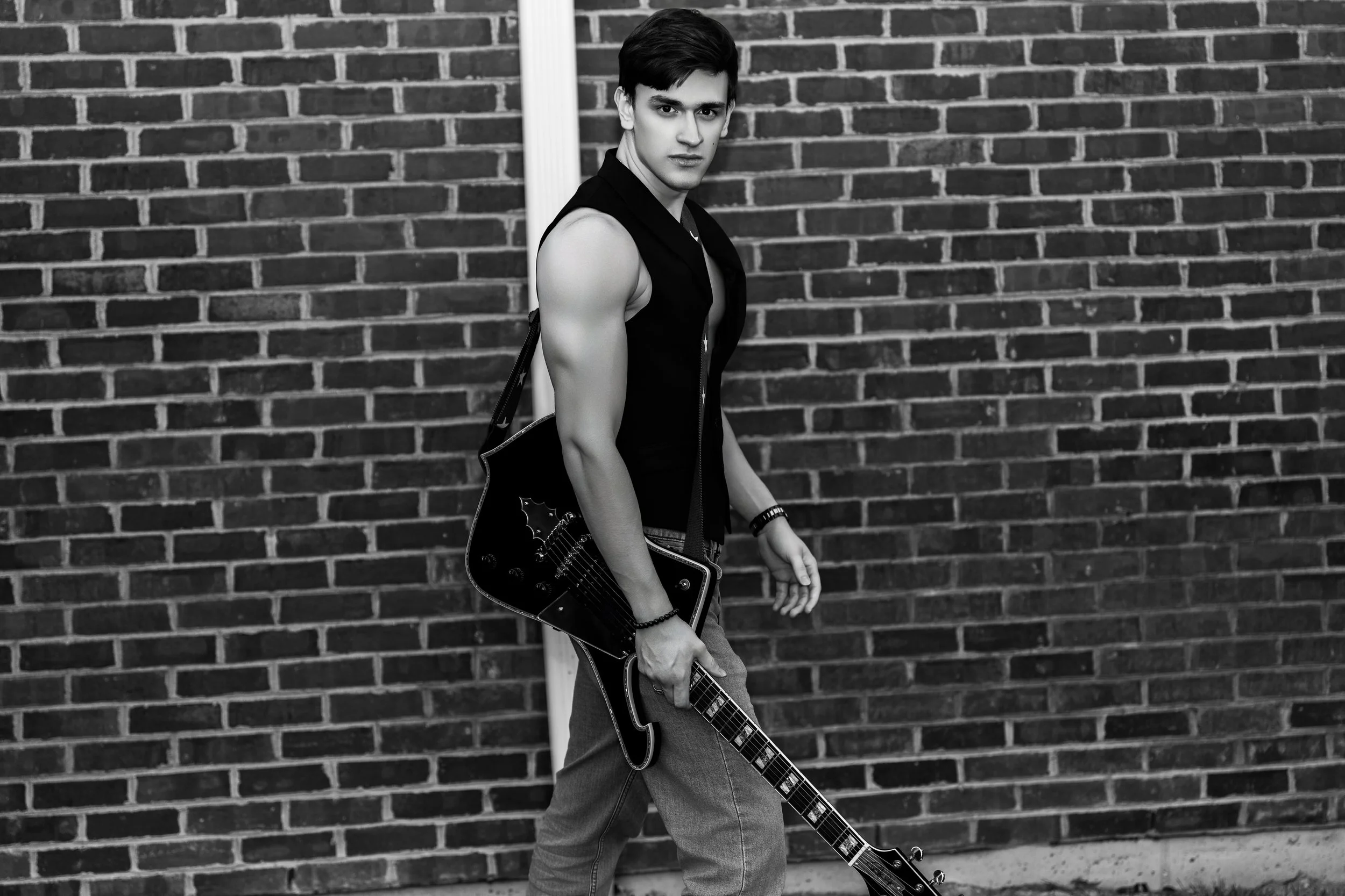 A young man standing against a brick wall, holding an electric guitar in his right hand, wearing a sleeveless vest, jeans, and accessories, looking at the camera.