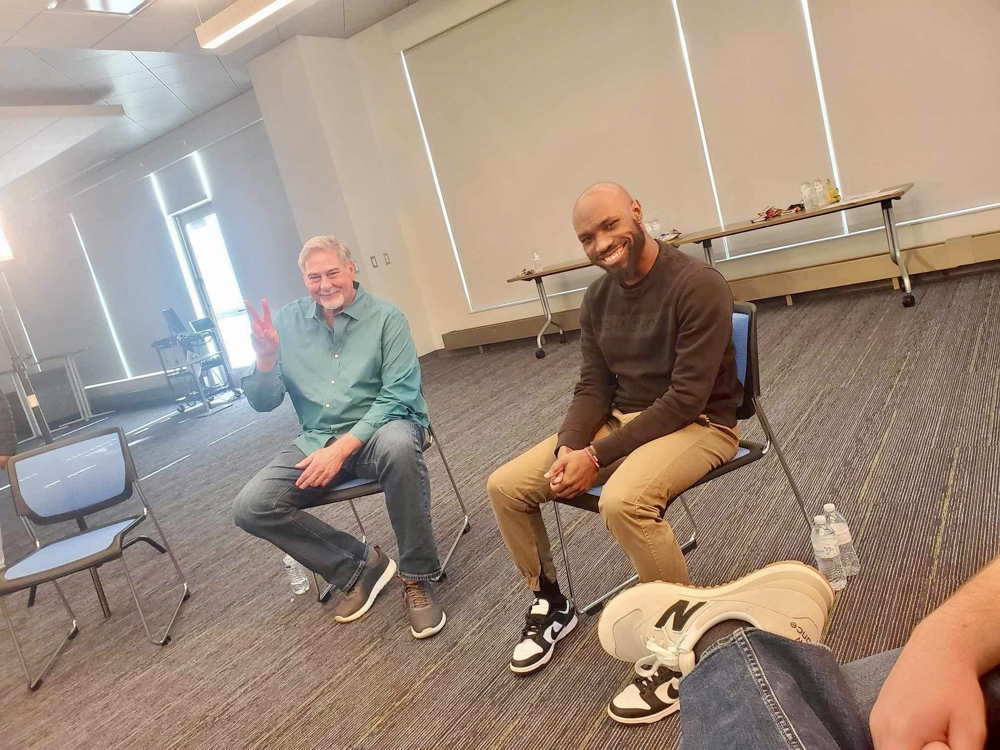 Two men sitting on chairs in a room, both smiling. The man on the left has gray hair and a beard, making a peace sign. The man on the right is bald with a beard, smiling. There are water bottles and a table with food and cups behind them.