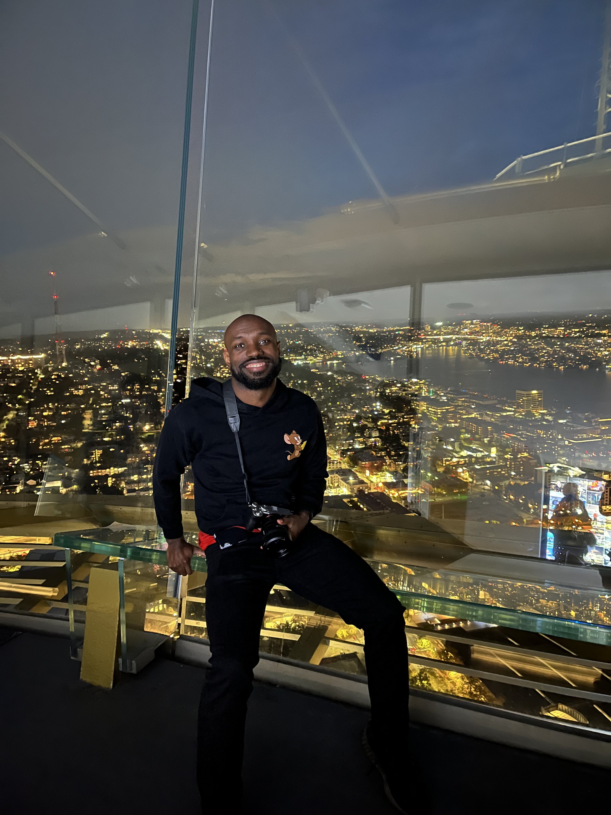 Man with camera posing in a glass observation deck overlooking a city skyline at night.