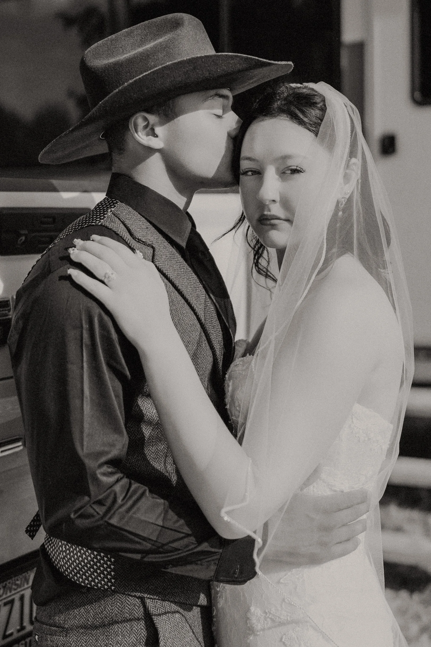 A black and white photo of a couple, with the man wearing a hat and suit, kissing the woman's forehead, who is dressed in a wedding gown with a veil.