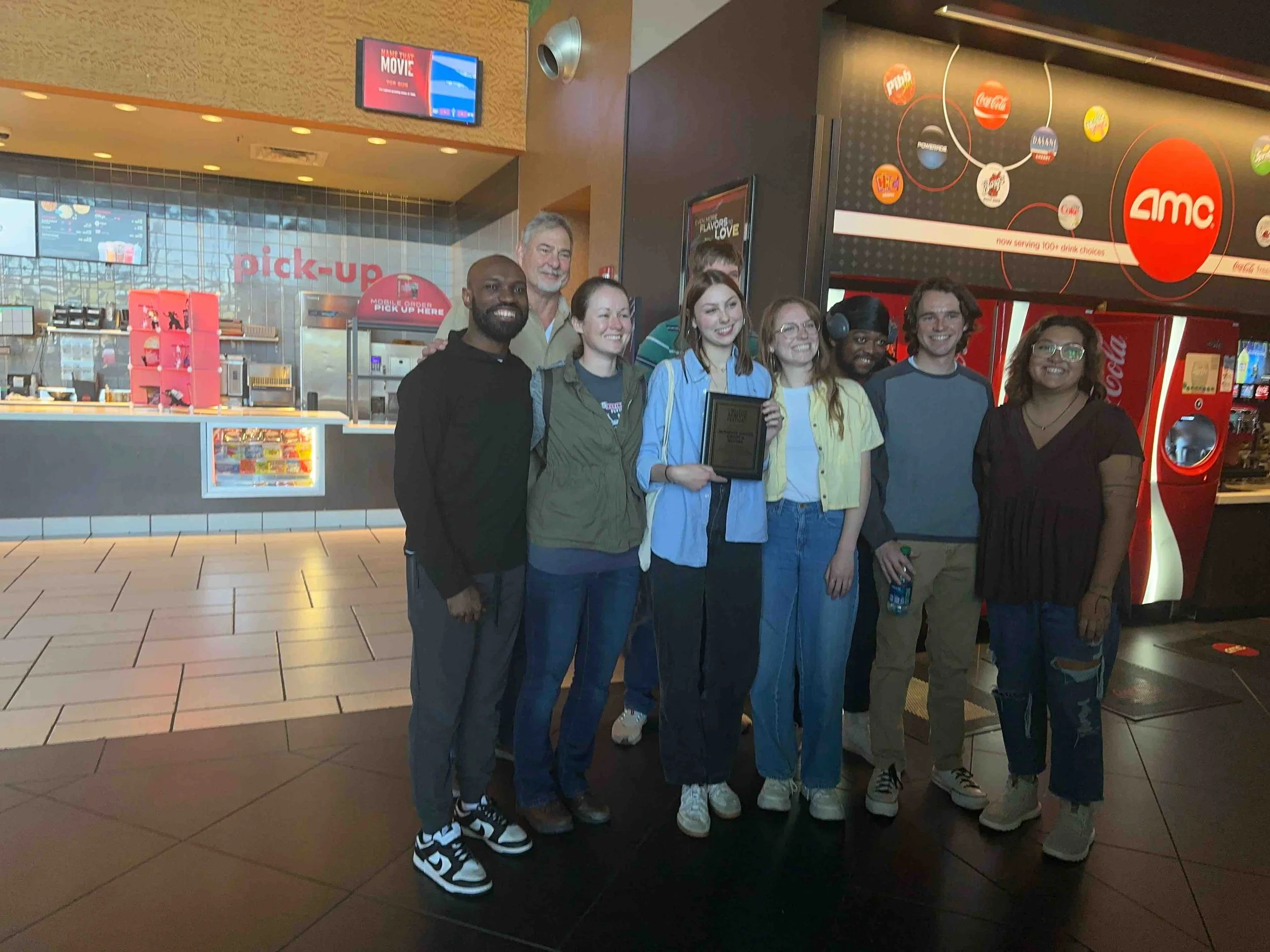 Group of nine people standing together in a movie theater lobby, some holding an award plaque, with a popcorn and drink counter in the background.