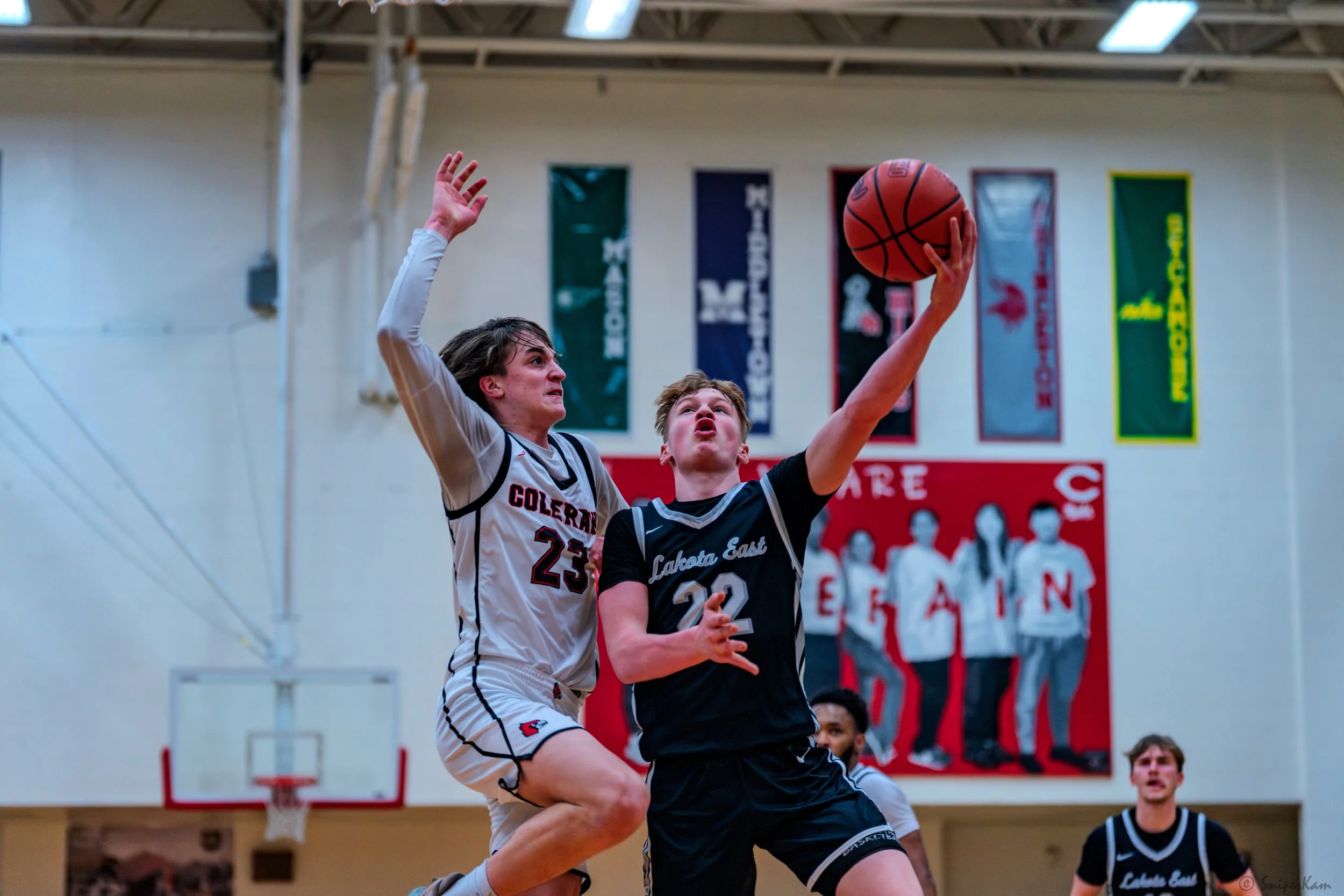 A basketball game with two male players mid-air near the basket, one from Colerain wearing jersey number 25 and the other from Lakota East wearing jersey number 22, competing for a rebound or shot attempt inside a gymnasium.