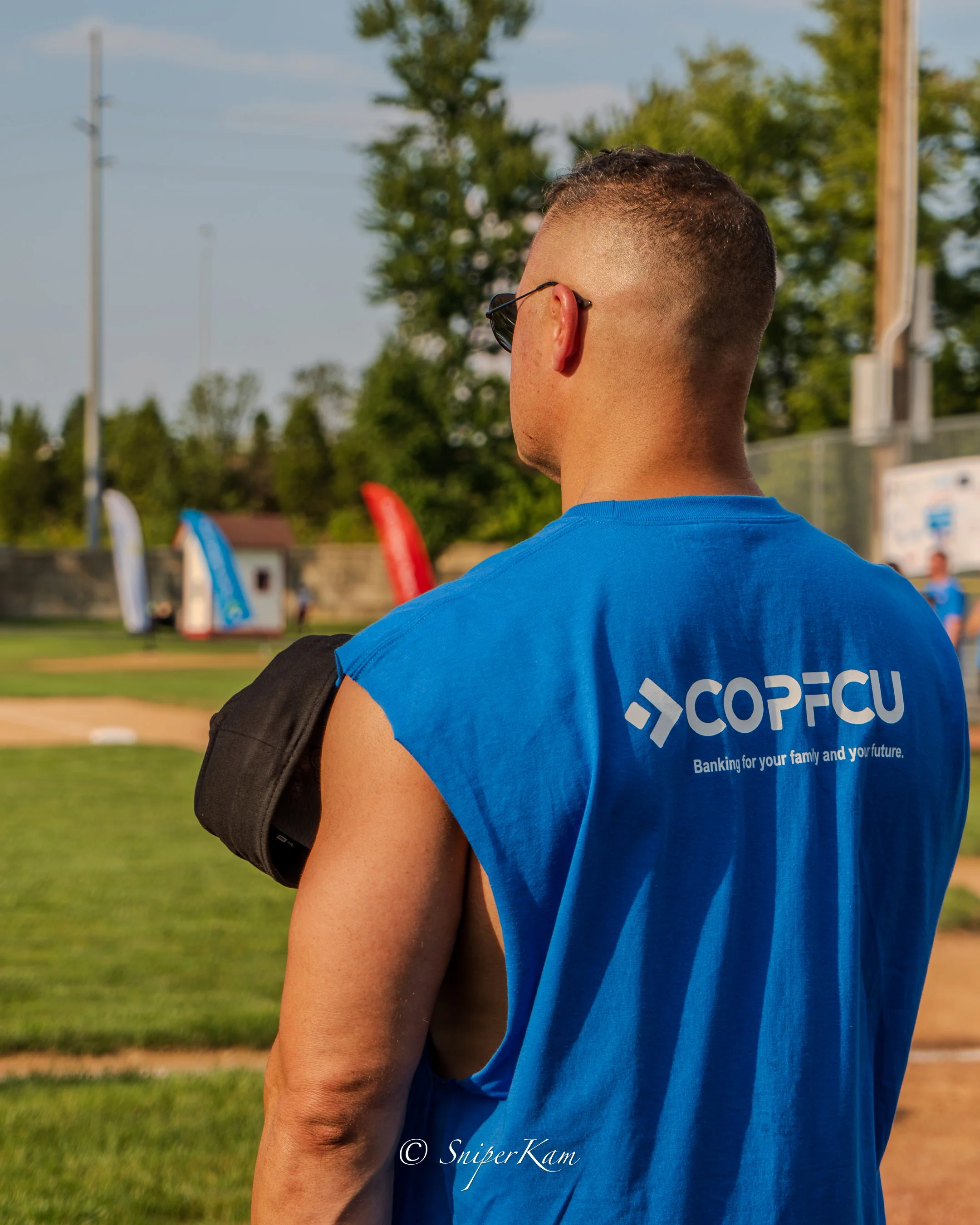 A man with a military-style haircut wearing sunglasses and a blue sleeveless t-shirt with the logo 'COPFCU' and the tagline 'Banking for your family and your future' stands on a sports field, facing away. He holds a black bag over his shoulder, and t