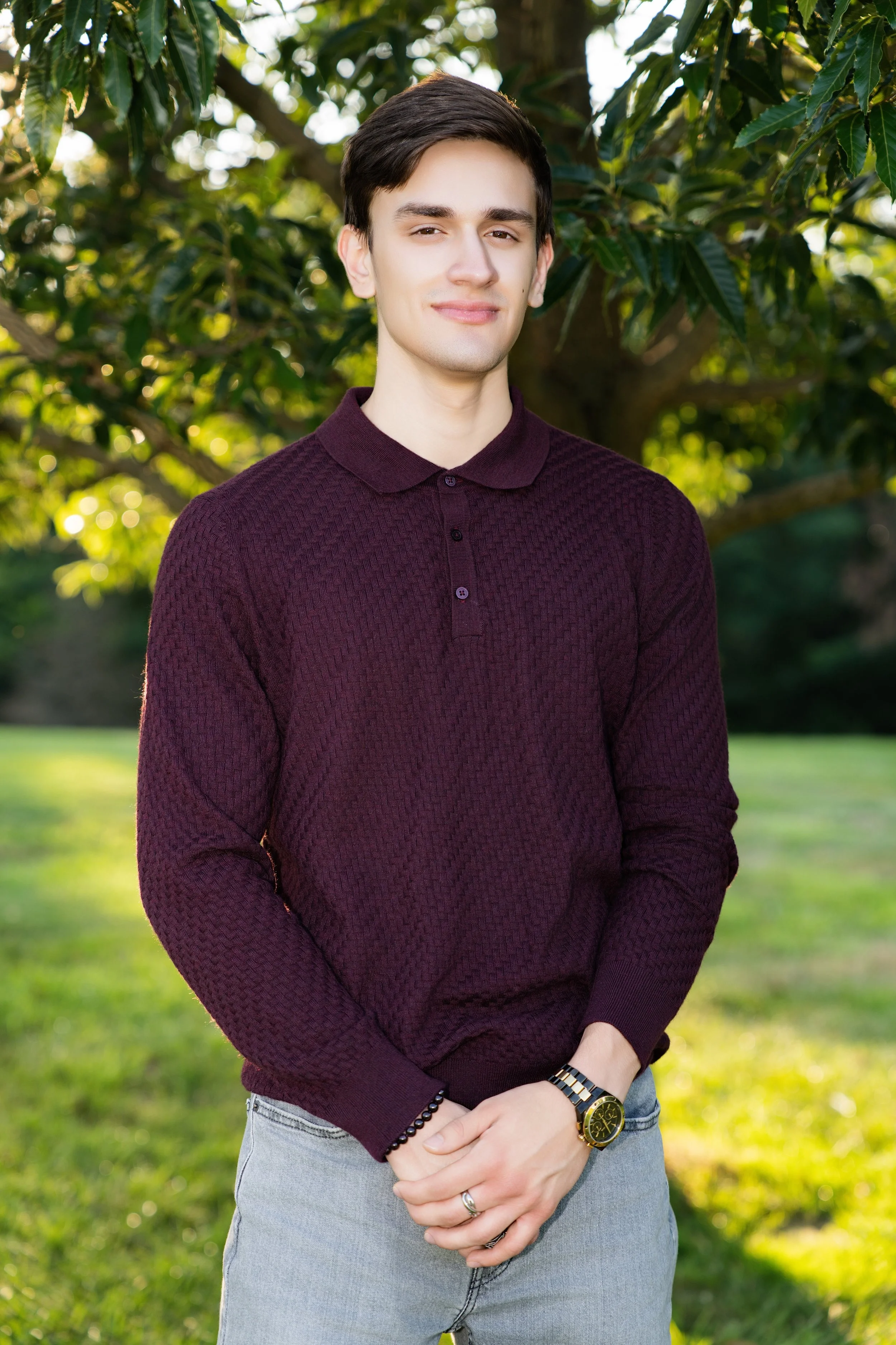 A young man standing outdoors in front of green trees, wearing a maroon textured sweater, light jeans, a watch, and a ring, with sunlight filtering through the leaves.