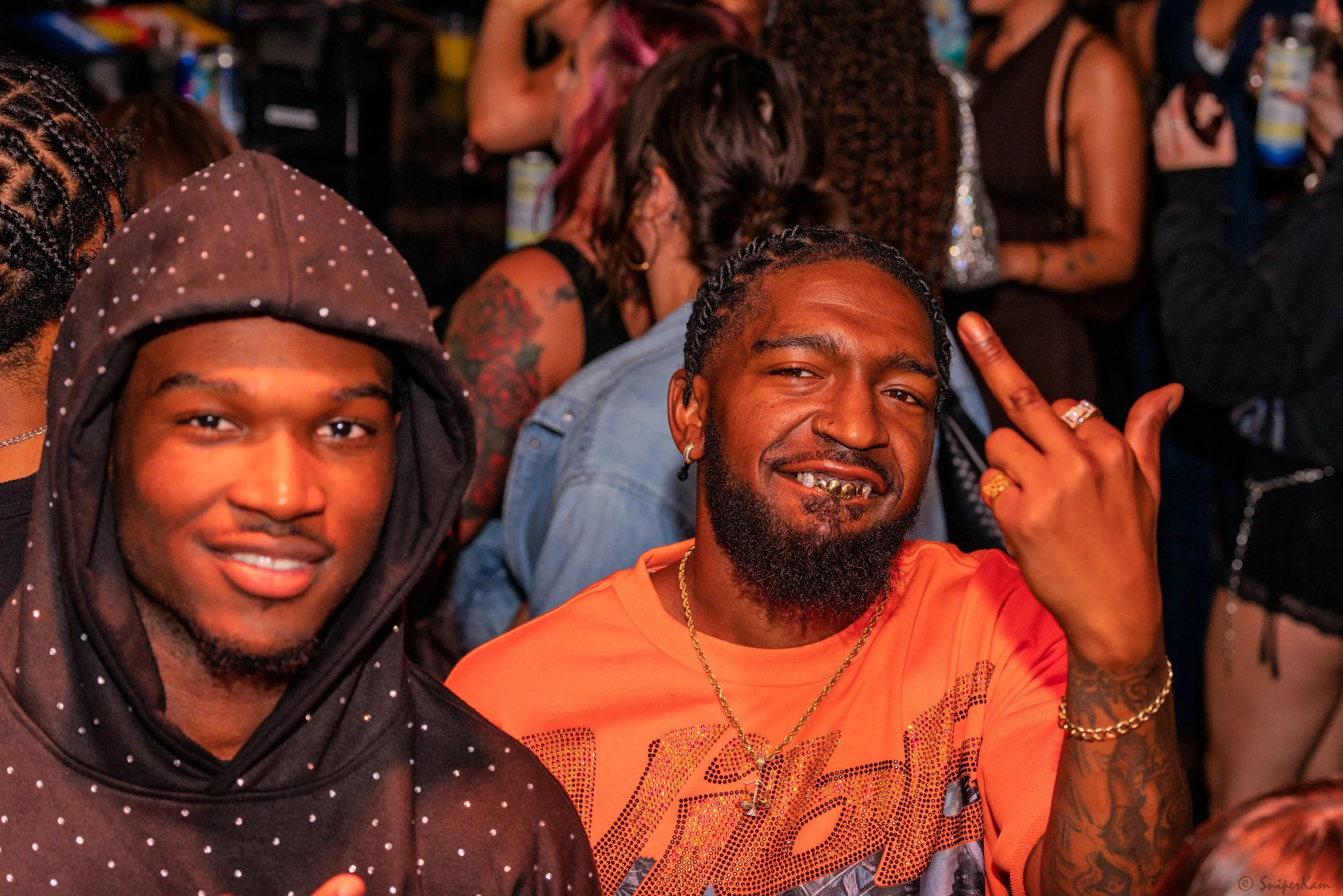 Two men at a crowded party or club, one wearing a dark hoodie with a hood decorated with white dots, the other in an orange shirt, making a rude hand gesture, with visible jewelry and facial piercings, surrounded by other partygoers.