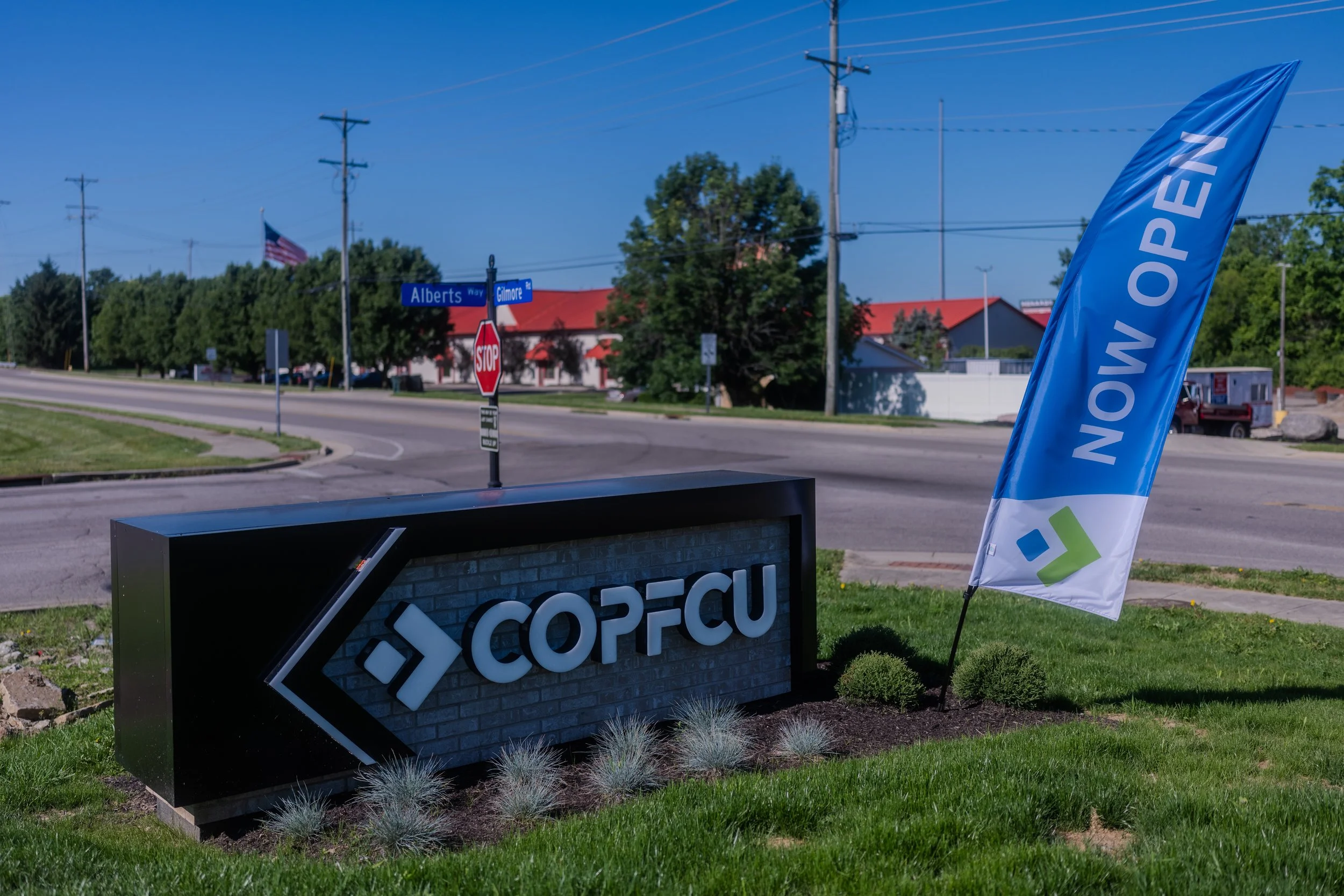 A blue flag with white text reading 'NOW OPEN' is displayed outdoors near a street intersection with street signs for Alberts Way and Gilmore. There is a black and white sign with reflective lettering in the foreground, and an American flag is visible in the background among green trees and buildings with red roofs.