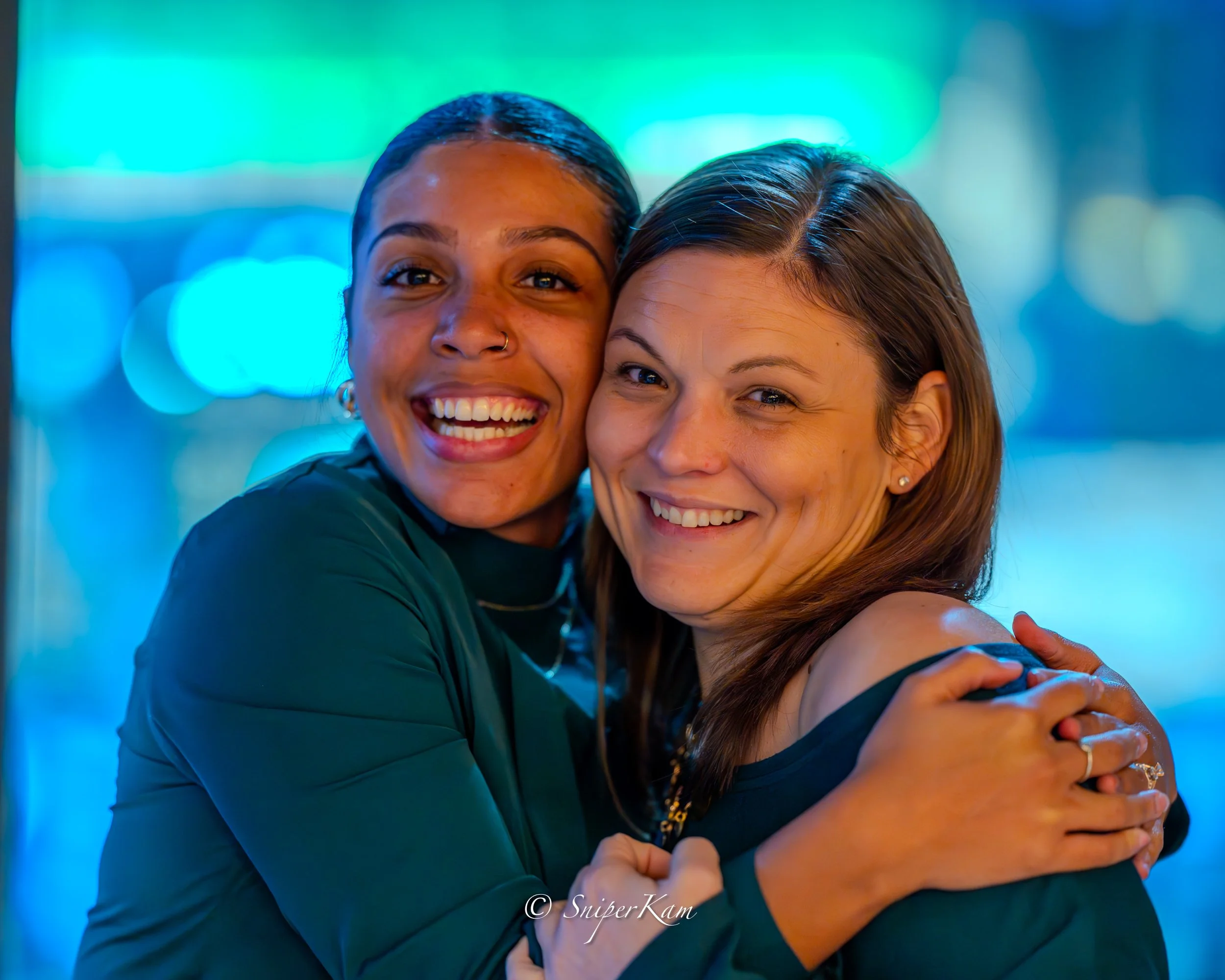 Two women hugging and smiling at the camera, with a colorful blurred city background at night.