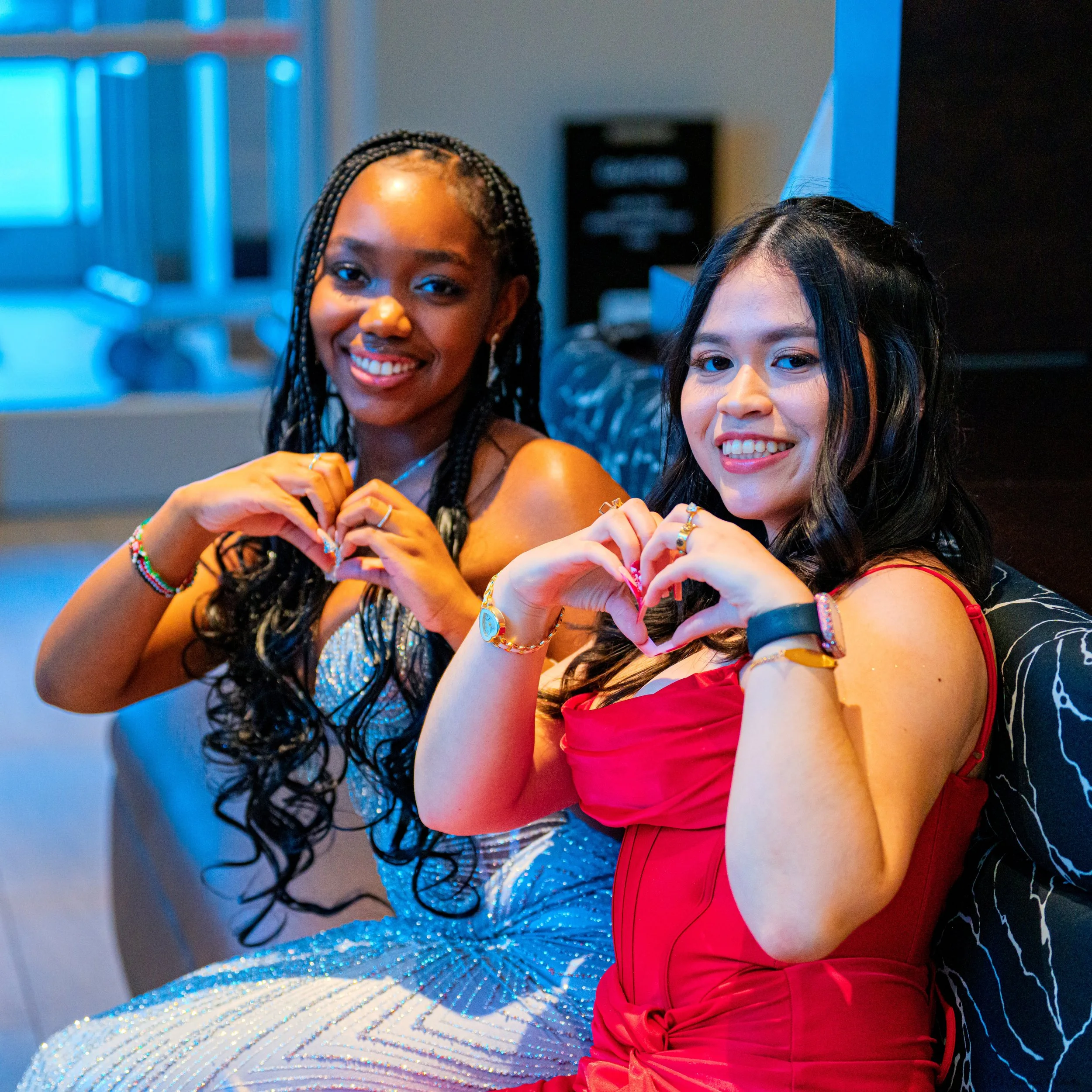 Two women sitting on a black and white patterned couch, smiling, and making heart shapes with their hands. One in a sparkly silver dress and the other in a red dress, both wearing jewelry and rings.