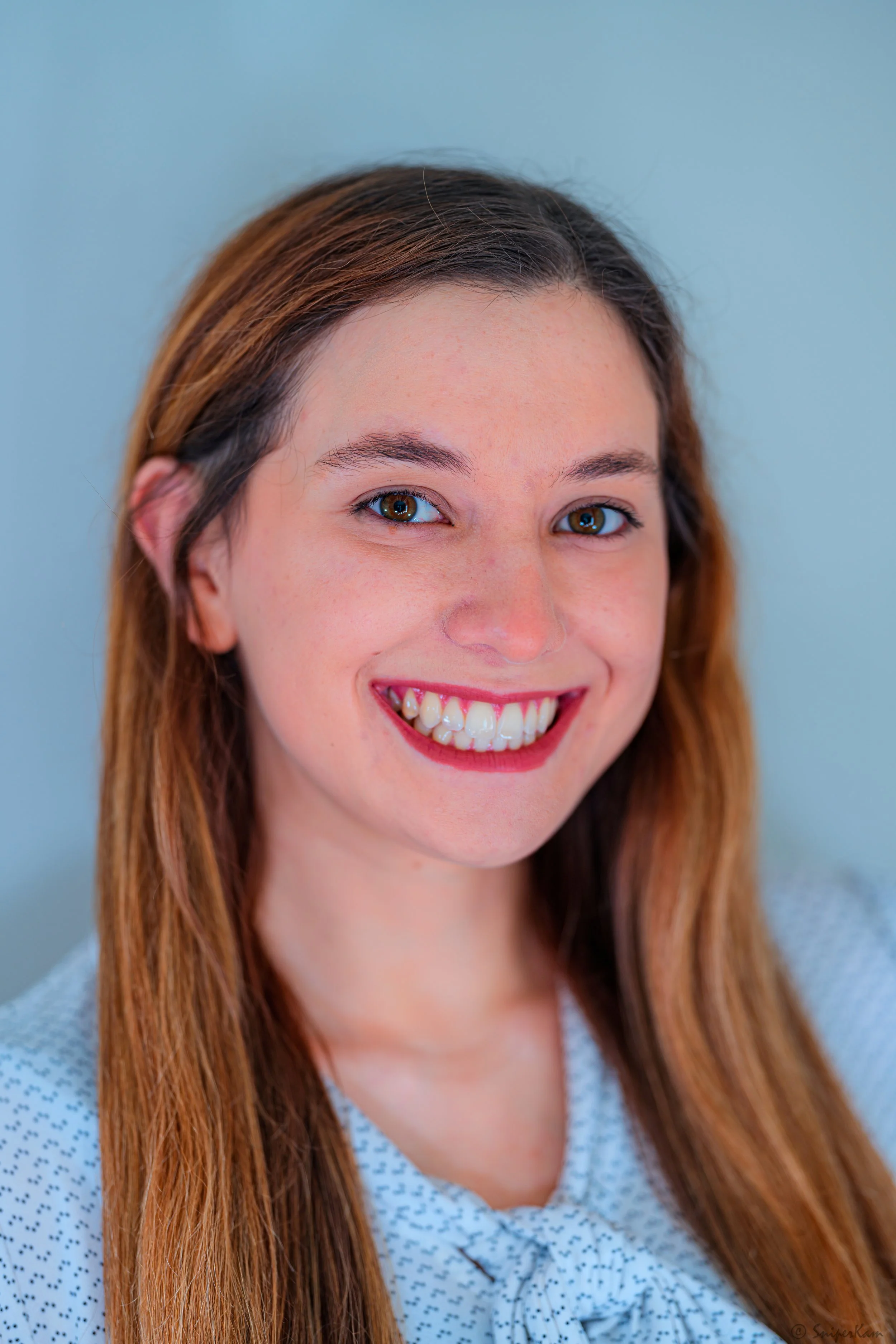 Close-up portrait of a young woman smiling with white teeth, long reddish-brown hair, and wearing a light-colored, patterned top against a light blue background.