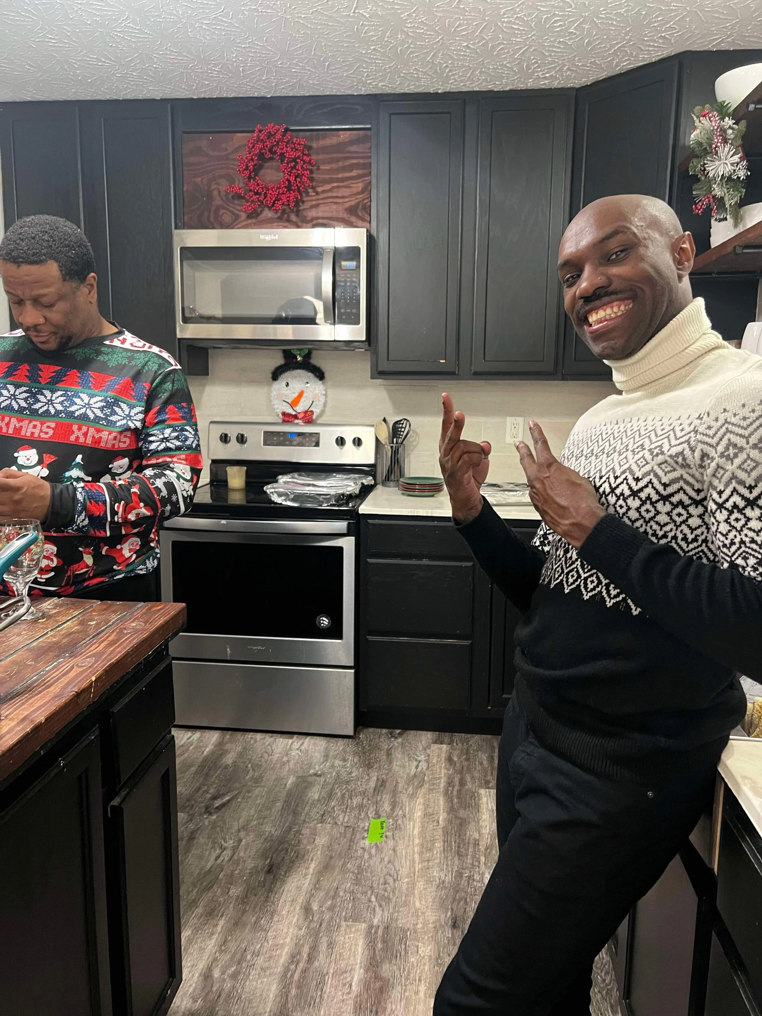 Two men in a kitchen decorated with Christmas decor. One man with a mustache is smiling and making a gesture with his hands, wearing a black and white sweater. The other man is looking down at something, wearing a colorful Christmas sweater.