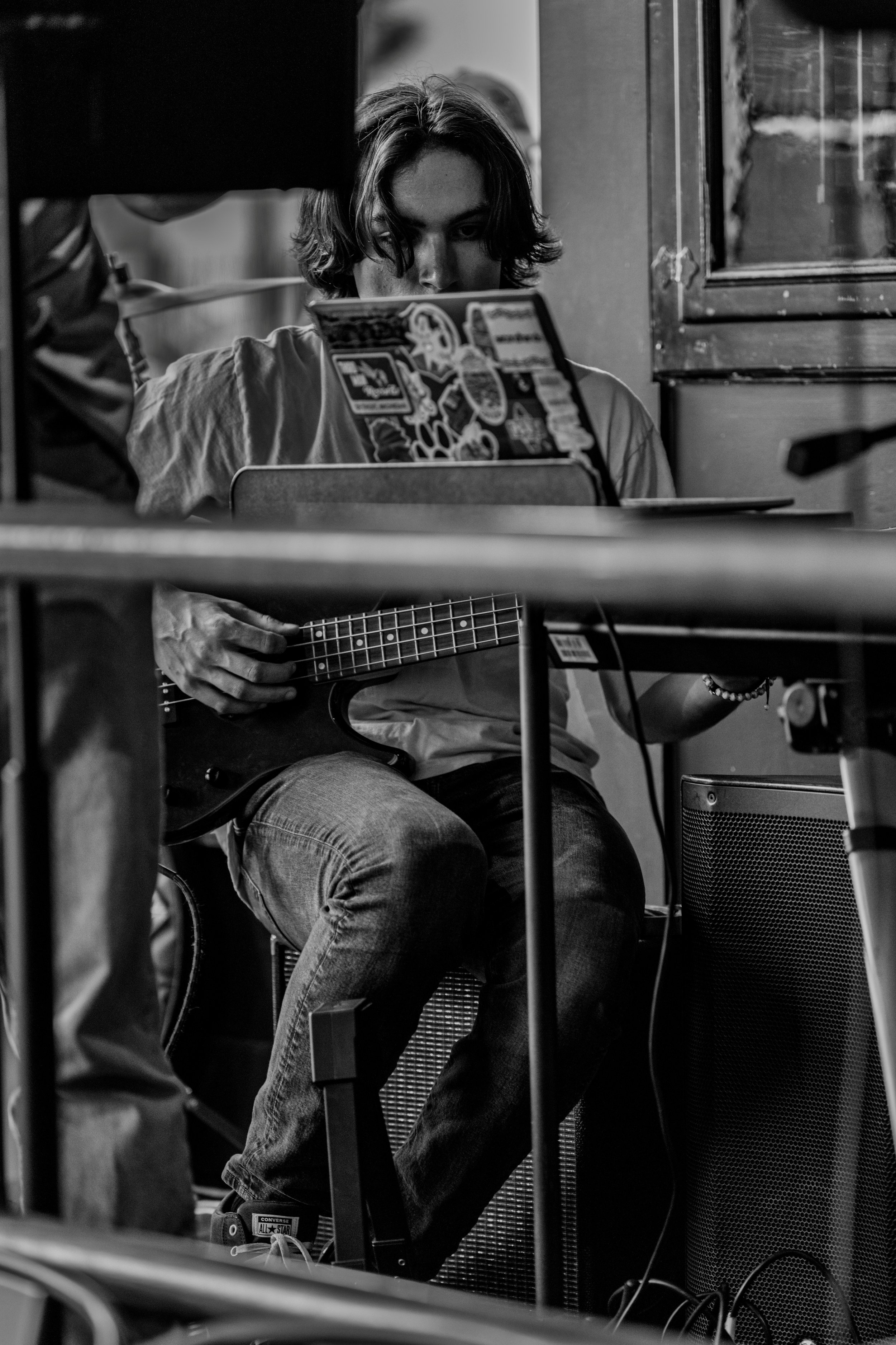 A young man with shoulder-length hair playing an electric guitar in a room, sitting near an amplifier and a window, with music sheets or sticker-covered sheet music stand in front of him, in black and white.