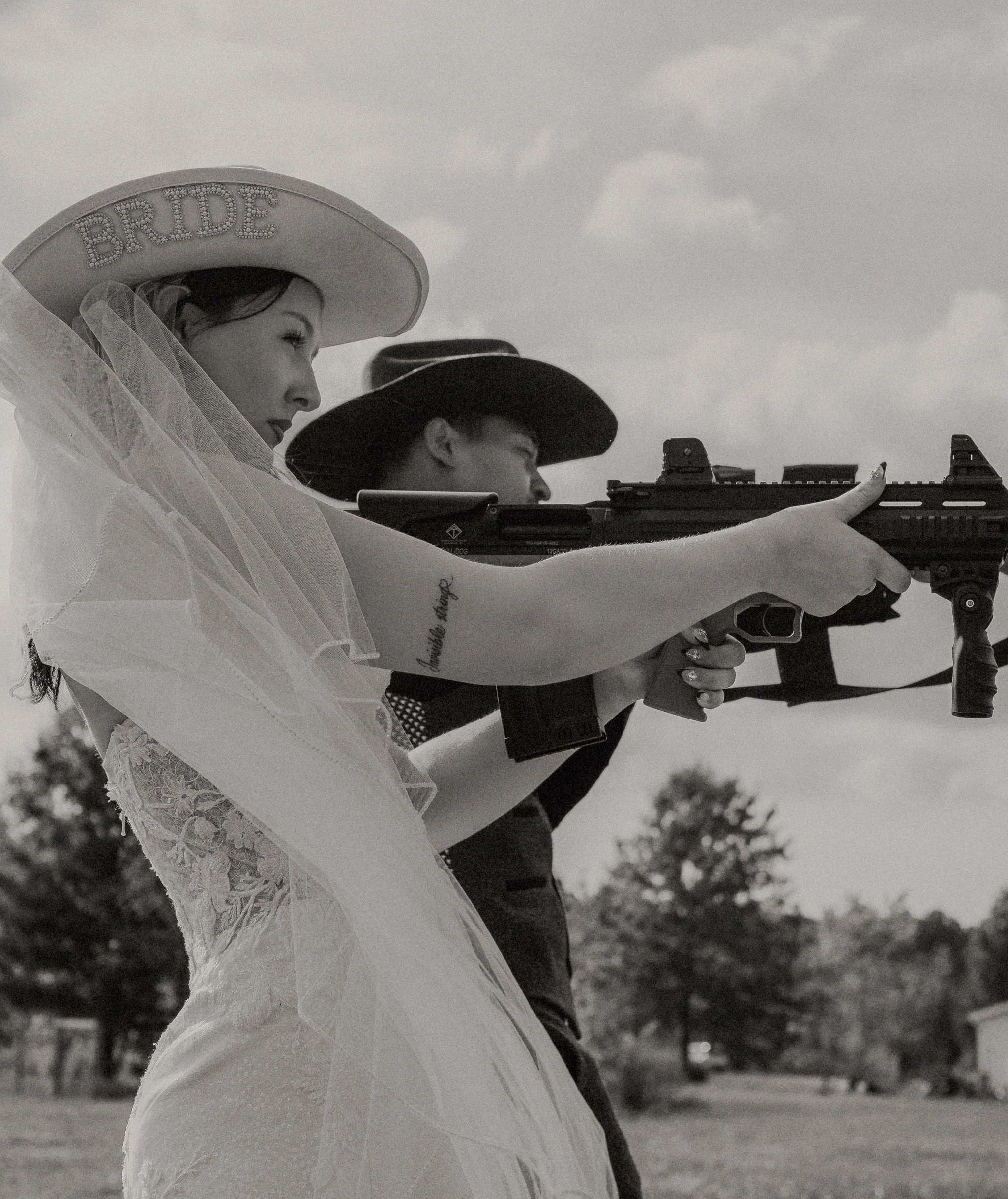 A woman wearing a hat with 'BRIDE' written on it and a veil, holding and aiming a rifle, standing outdoors with trees in the background.