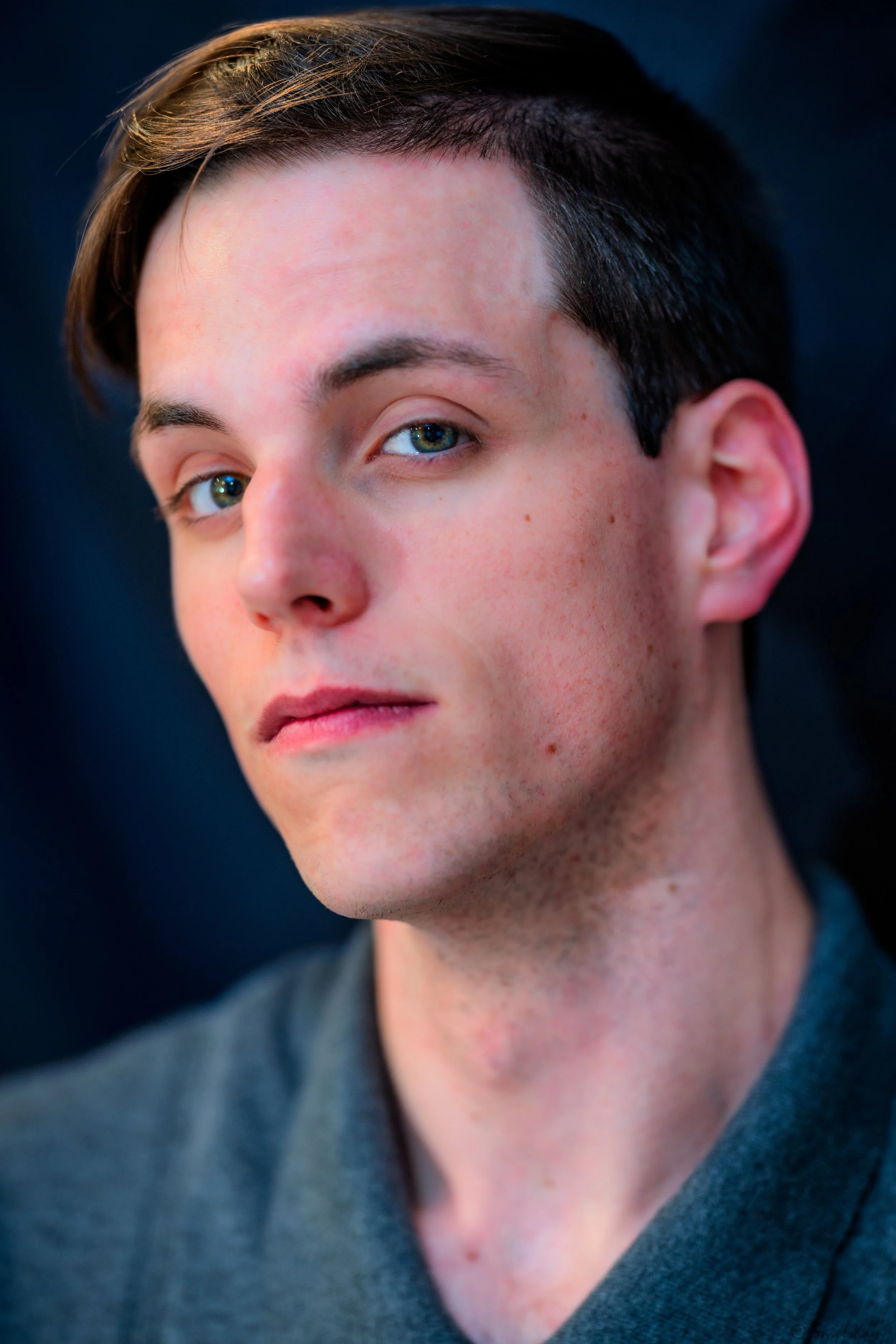 Close-up of a young man with fair skin, blue eyes, and dark brown hair, looking confidently at the camera with a serious expression.