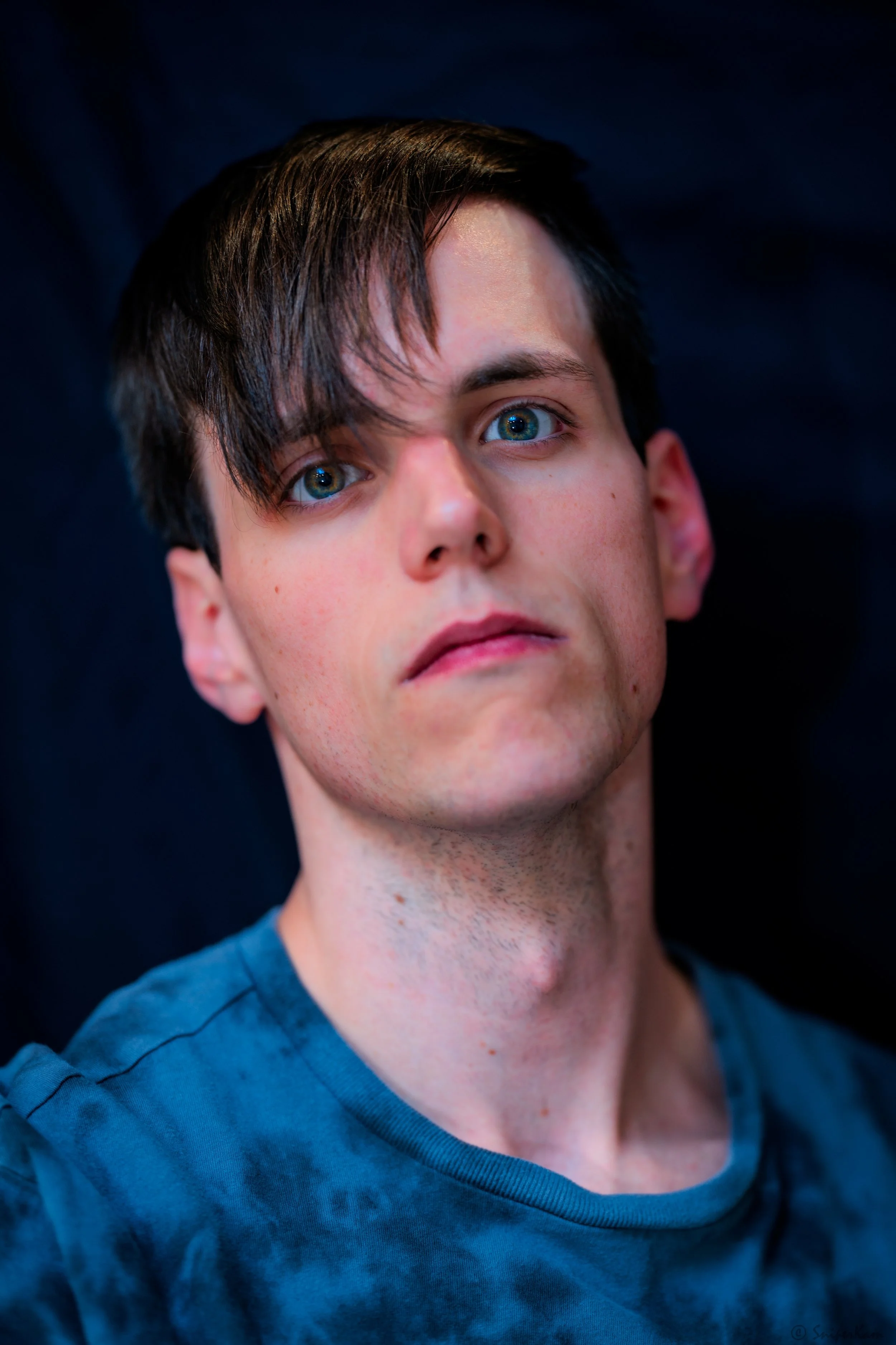 Close-up portrait of a young man with dark hair, blue eyes, and fair skin, wearing a blue tie-dye shirt, looking directly at the camera against a dark background.