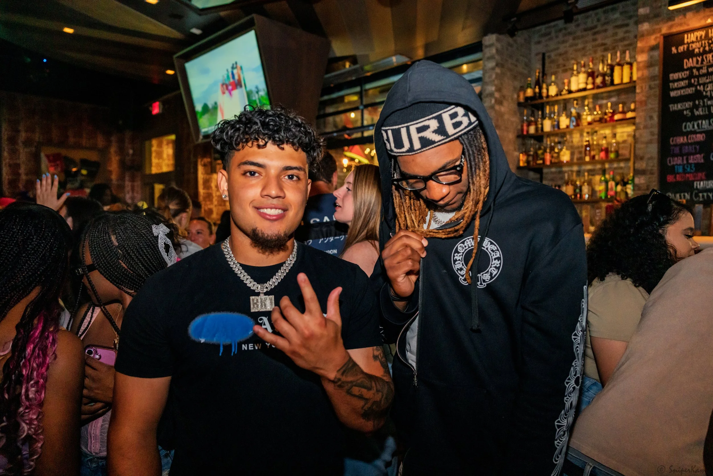 Two young men posing together in a lively bar or club with other people around and a bar shelf with bottles in the background.