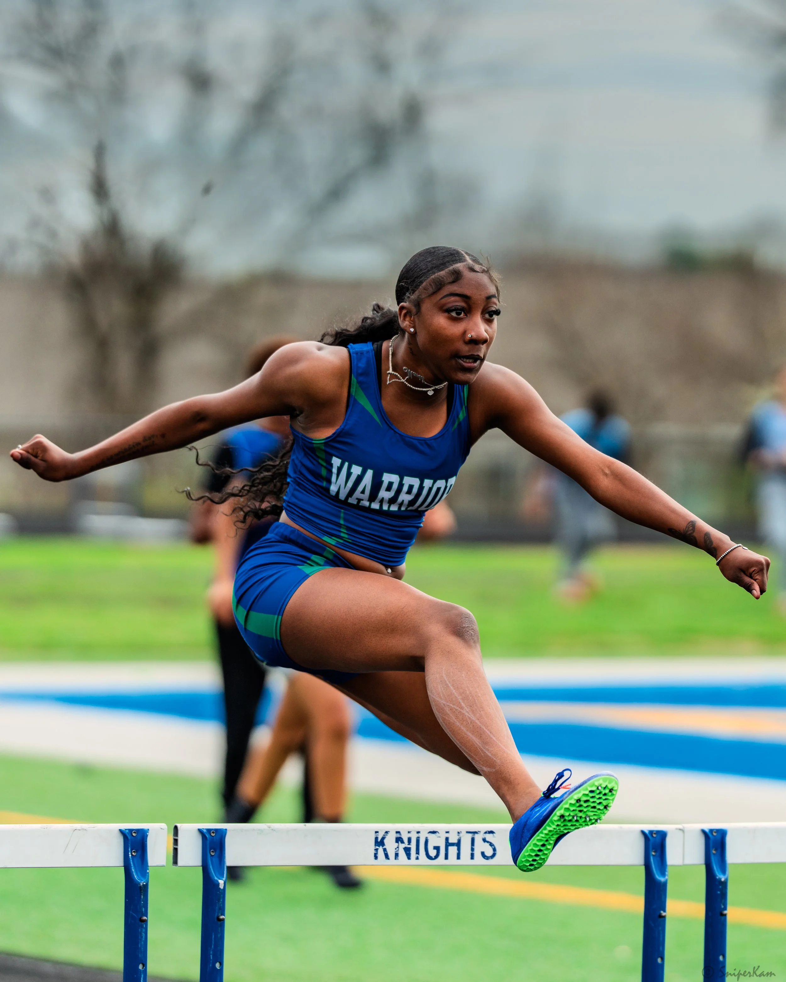 A female athlete in a blue uniform with the word 'WARRIORS' on it is jumping over a hurdle labeled 'KNIGHTS' during a track and field event.