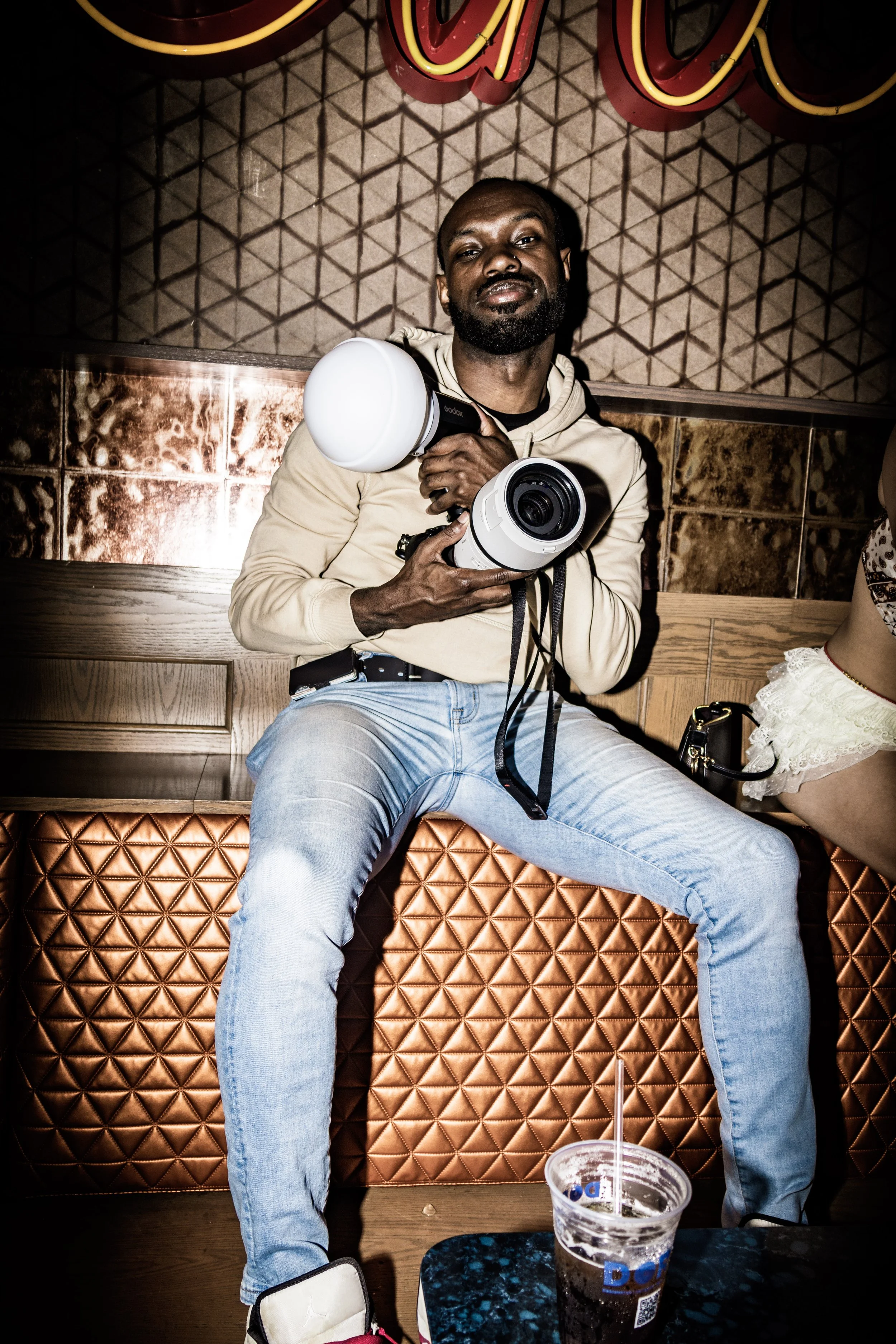 A man holding a professional camera sits on a bench in a bar or cafe, with a drink in front of him, and a woman partially visible beside him.