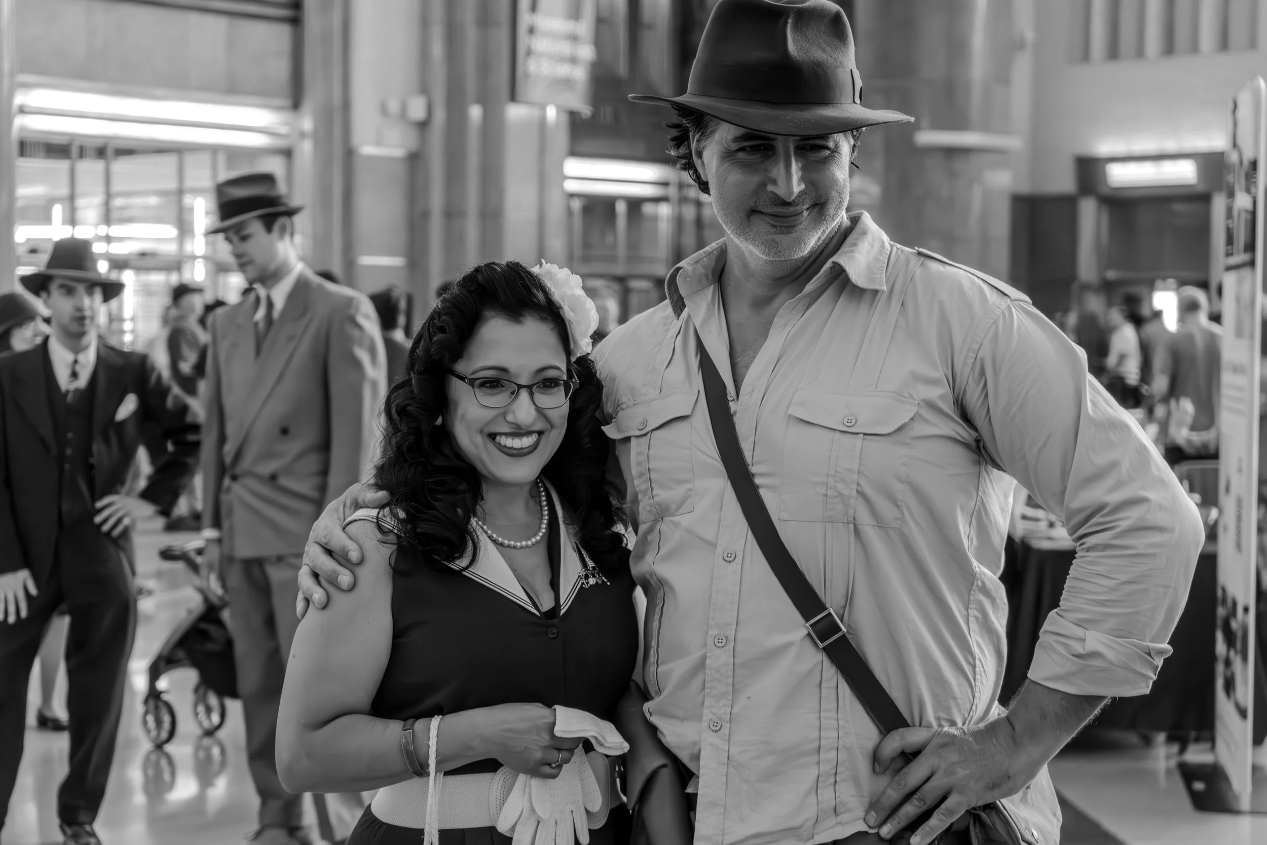 A woman in vintage clothing and glasses smiling next to a man in a hat and casual shirt, both posing for a photo, with other vintage-dressed people in the background.