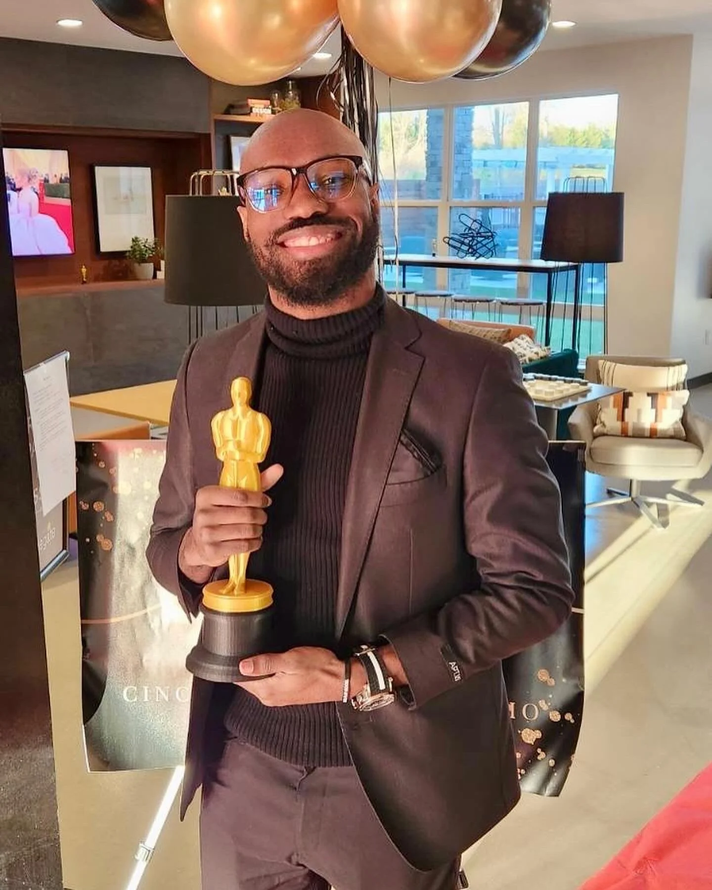 A man in a dark suit and glasses smiling and holding an Oscar trophy, standing in a well-lit room with balloons, a poster, and modern furniture.