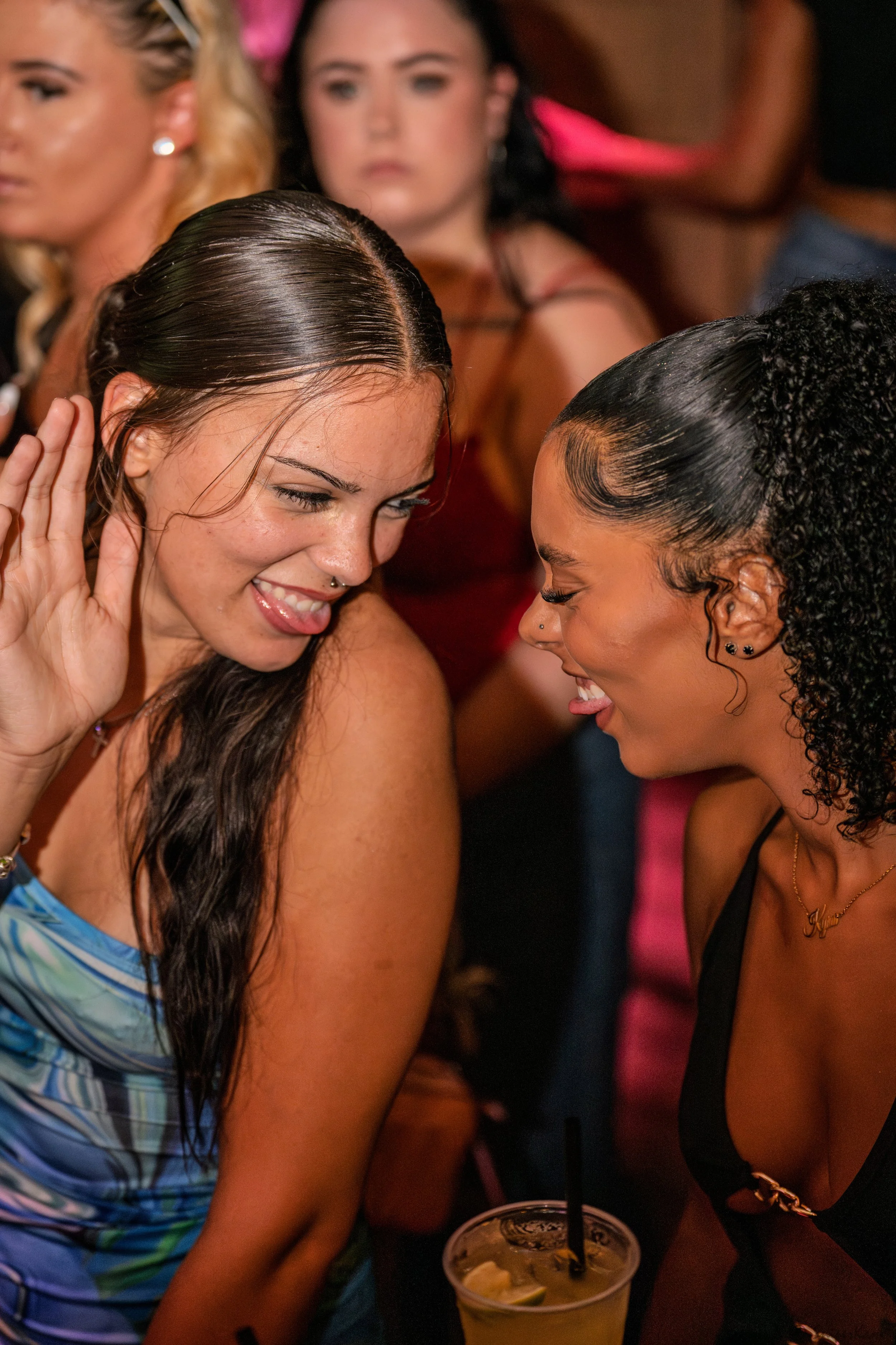 Two women are smiling and laughing close together at an indoor party or bar, with other women in the background. One woman has long dark hair and is wearing a strapless top, while the other has curly black hair styled in a bun and is wearing a black 