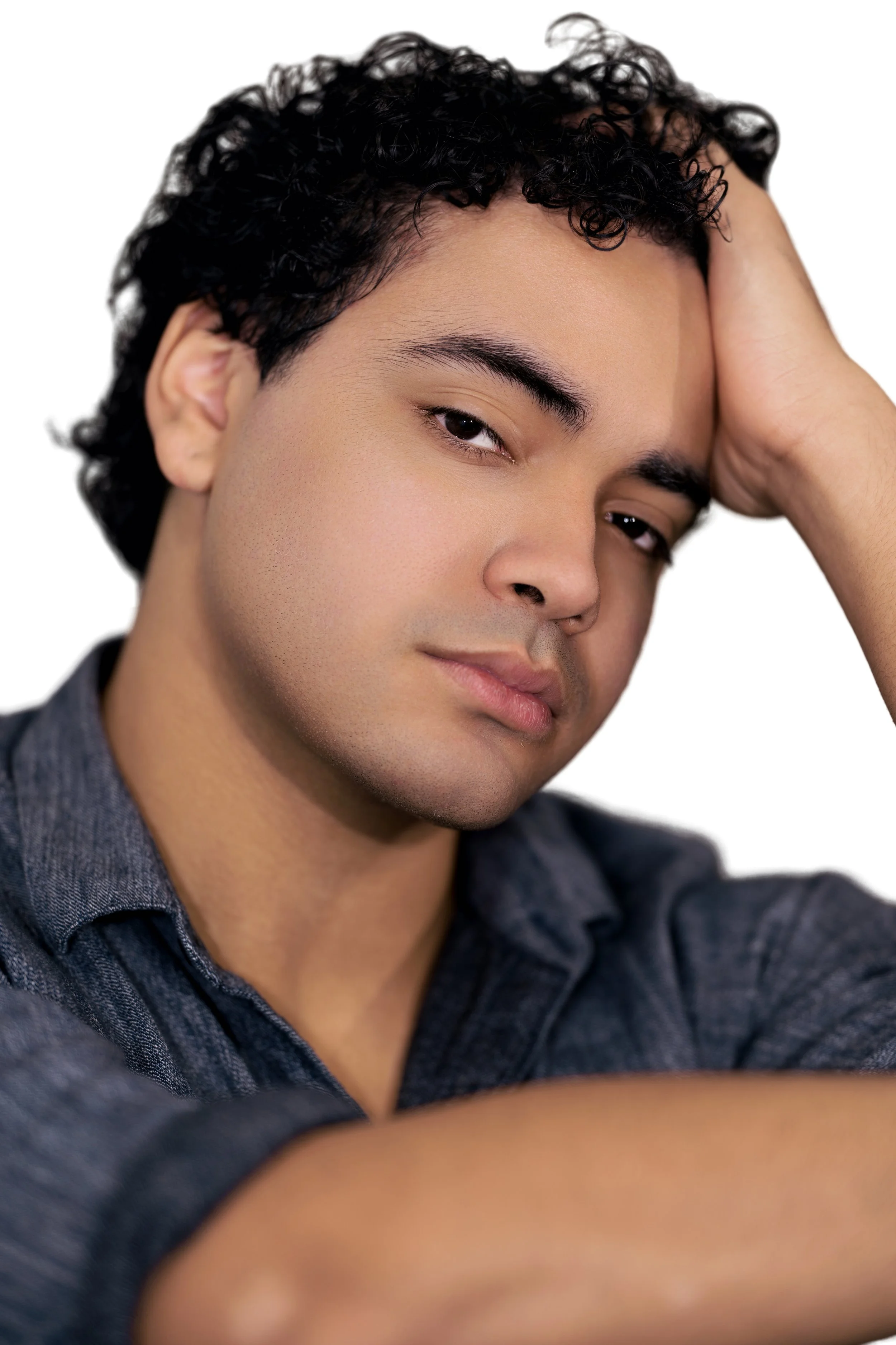 Close-up of a man with dark curly hair, resting his head on his hand, wearing a dark denim shirt, looking confidently at the camera.