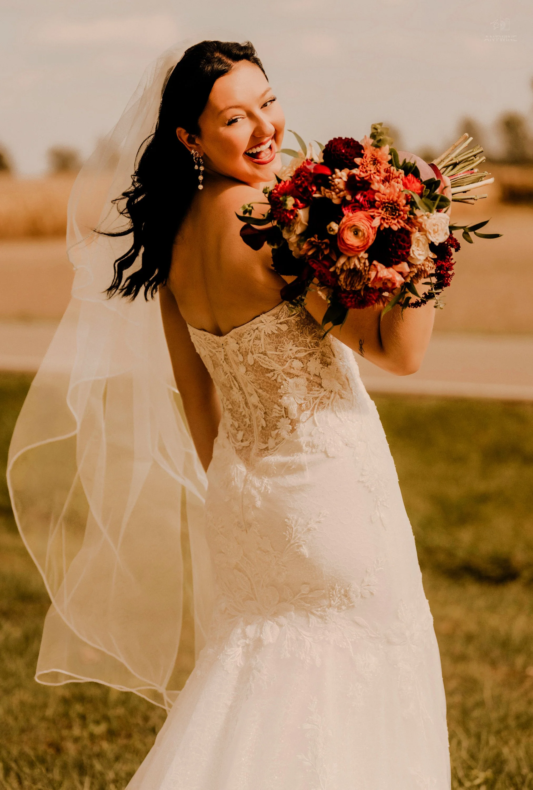 A smiling bride in a wedding dress holding a bouquet of red, pink, and white flowers outdoors at sunset.