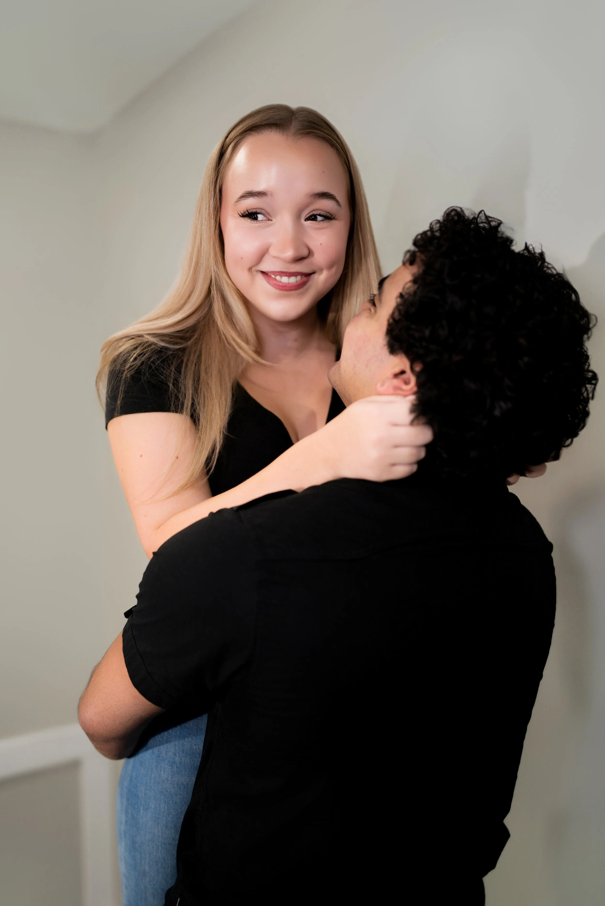 A young woman with blonde hair smiles as a man with curly black hair holds her. They are close together, touching faces, in a friendly or romantic moment indoors.