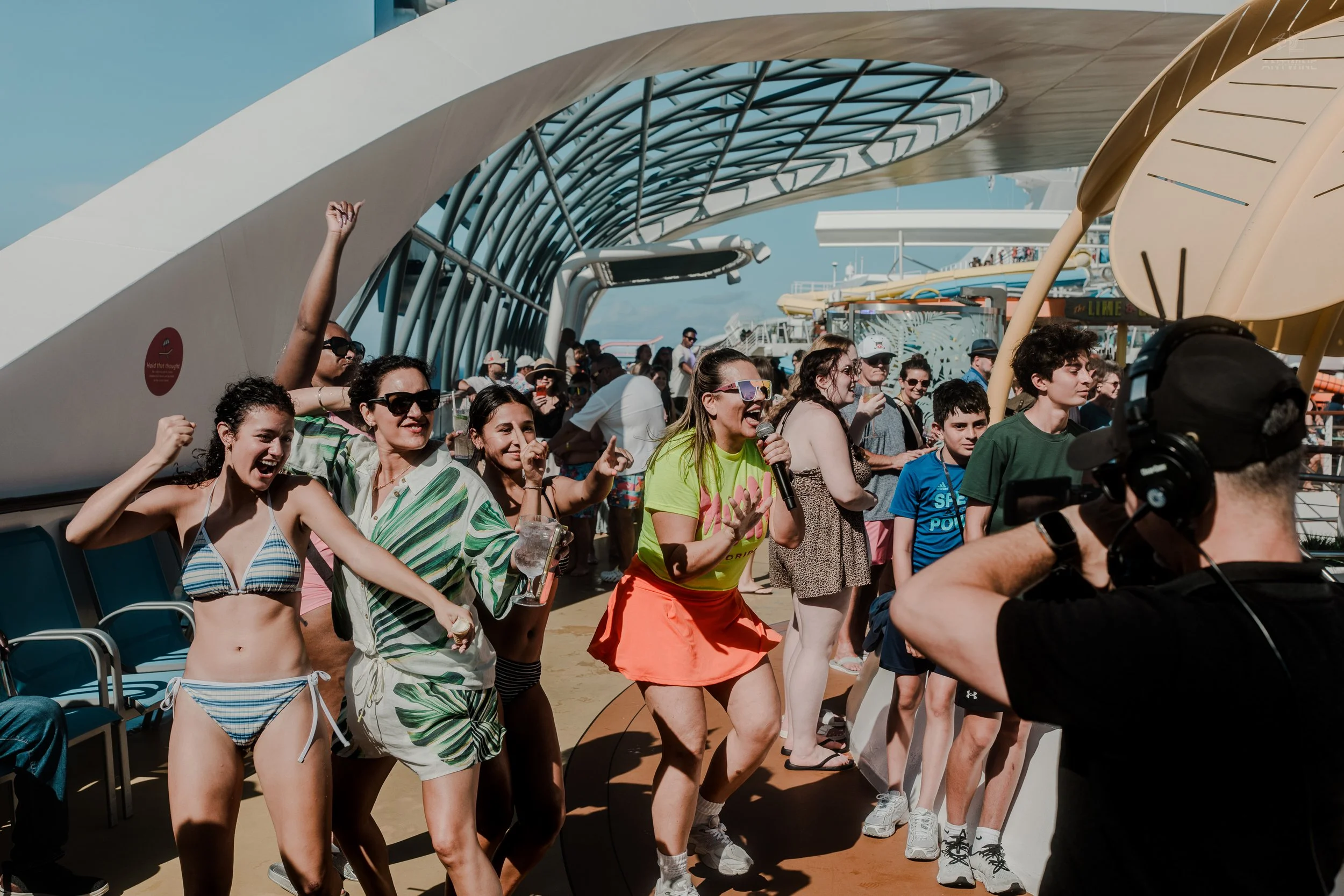 People aboard a cruise ship deck, with some dancing and others taking photos, under a modern architectural structure.