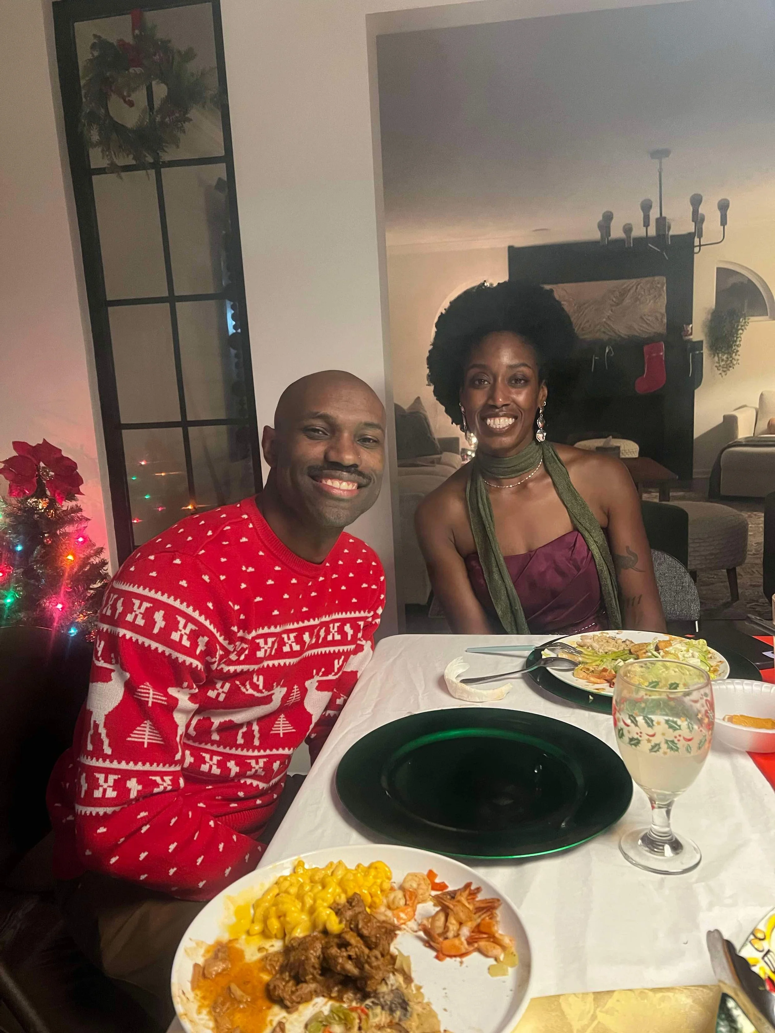 A man and woman smiling and sitting at a holiday dinner table with plates of food, a glass of white wine, and a Christmas tree in the background.